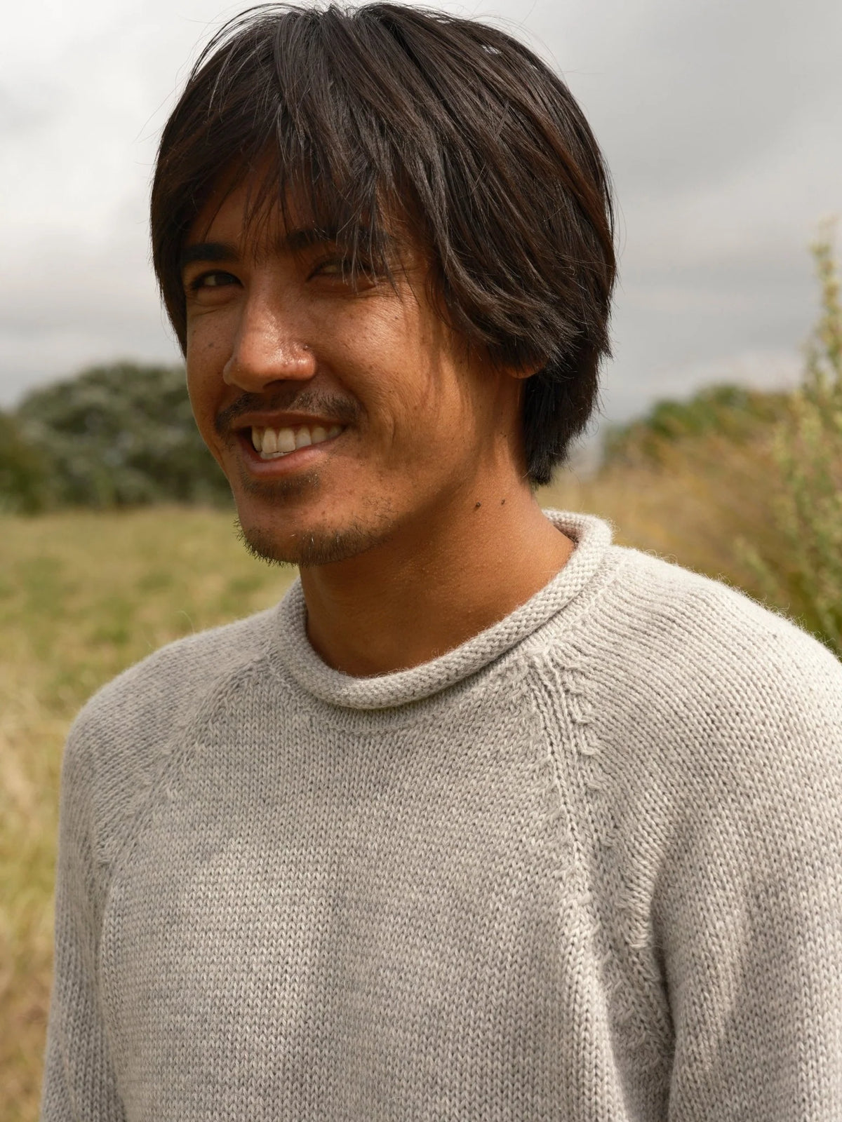 Close smiling portrait of a man wearing the Mollusk Fisherman Sweater in Pearl Gray outdoors, showing the rolled crew neckline and heathered pearl gray alpaca blend knit with subtle texture, against a blurred golden coastal meadow under an overcast sky
