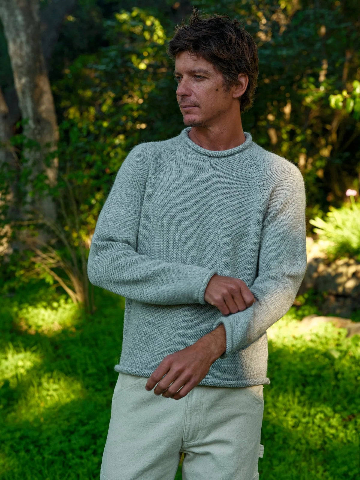 Three-quarter front view of a man with arms crossed wearing the Mollusk Fisherman Sweater in Pearl Gray, showing the relaxed crew neck, raglan sleeves, rolled cuffs, and heathered light gray alpaca blend knit, styled with cream pants in a lush sunlit garden
