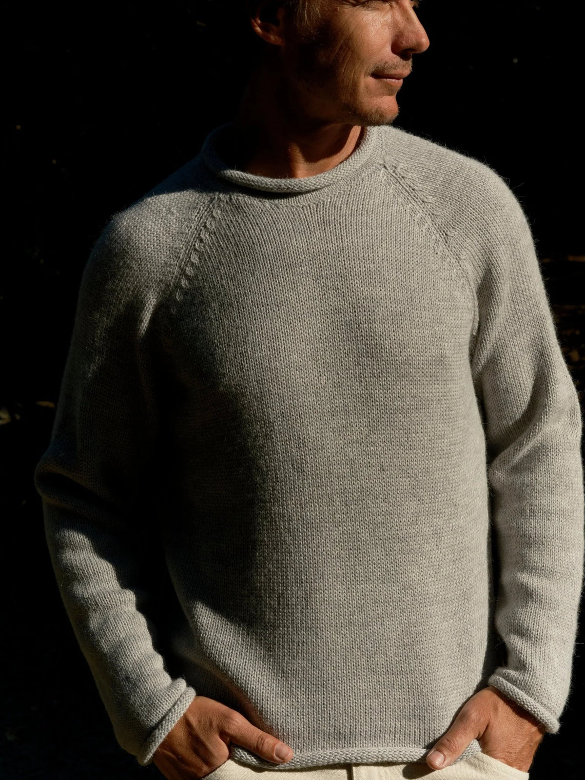 Three-quarter view of a man looking to the side wearing the Mollusk Fisherman Sweater in Pearl Gray, showing the relaxed fit, raglan sleeve seam, rolled crew neck, and heathered light gray alpaca blend knit, styled with cream pants in a sunlit garden with green foliage
