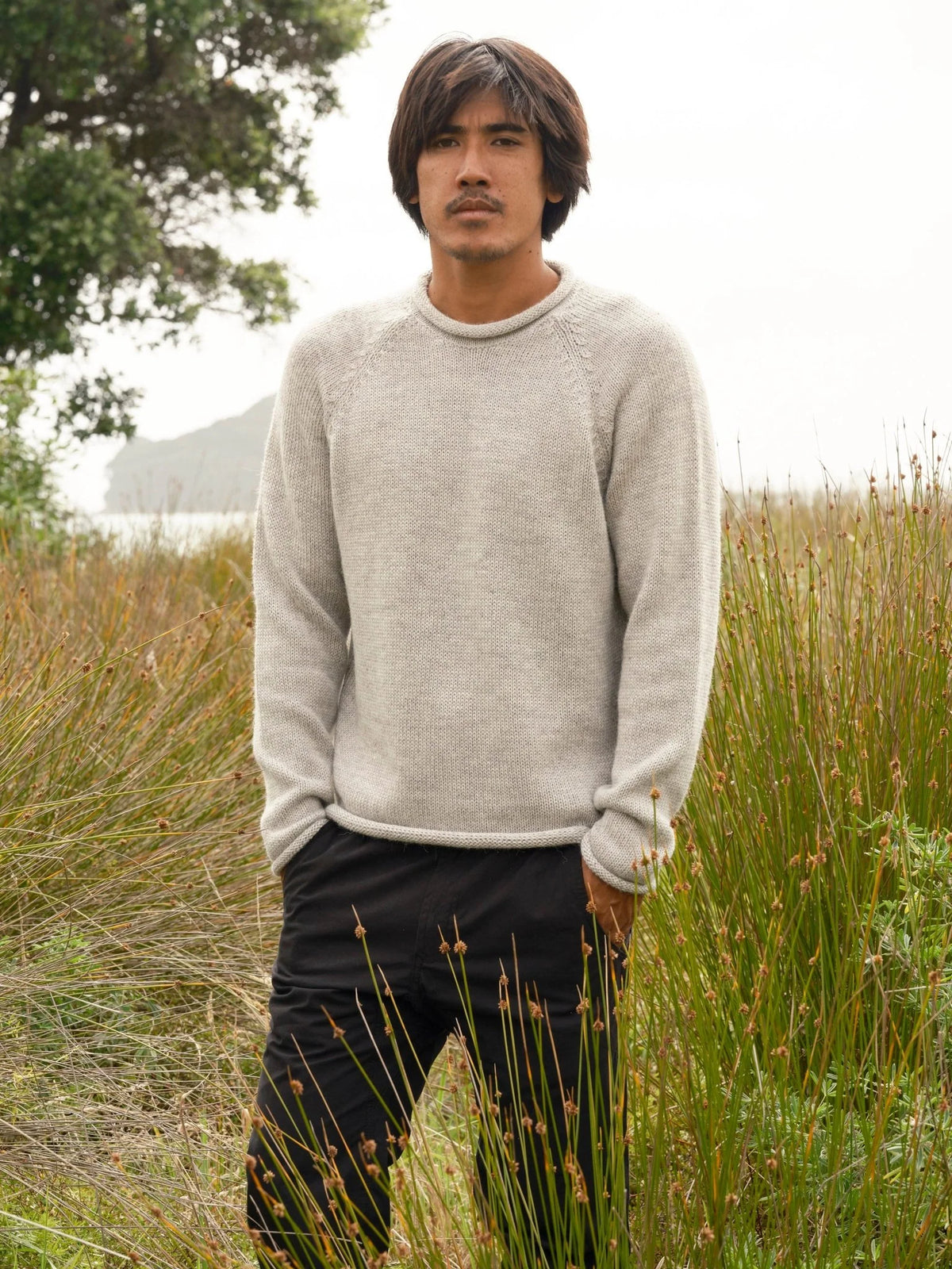 Front view of a man standing in a coastal meadow wearing the Mollusk Surf Shop Fisherman Sweater in Pearl Gray, showing the relaxed crew neck, raglan sleeves, rolled hem, and heathered light gray alpaca blend knit, styled with black pants against tall dry grasses and a misty ocean backdrop
