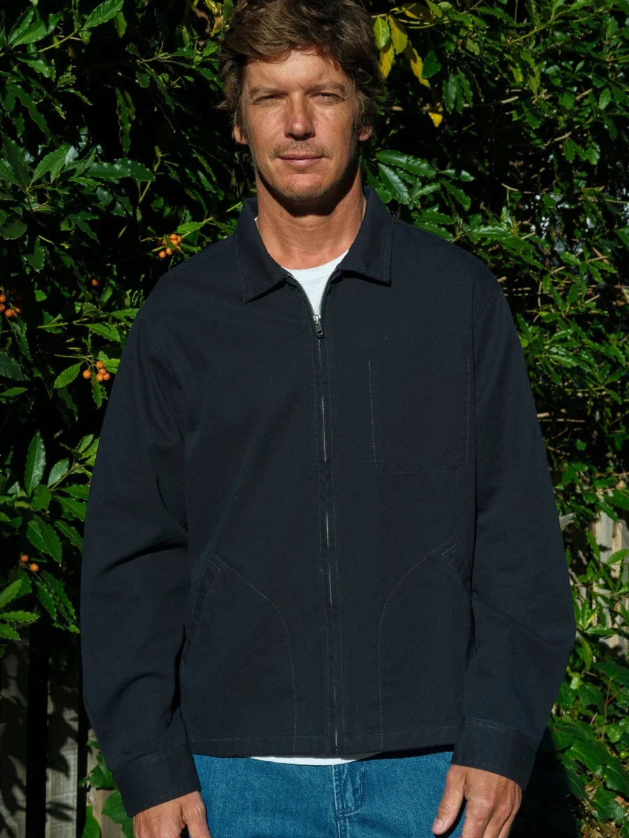Front view of a man wearing the Mollusk Surf Shop Gas Station Jacket in Navy zipped closed, showing the spread collar, full-length zipper, curved welt hand pockets, and relaxed fit in navy 100% cotton twill, styled with a white tee and bright blue jeans against a sun-dappled garden hedge
