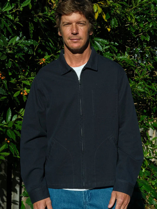 Front view of a man wearing the Mollusk Surf Shop Gas Station Jacket in Navy zipped closed, showing the spread collar, full-length zipper, curved welt hand pockets, and relaxed fit in navy 100% cotton twill, styled with a white tee and bright blue jeans against a sun-dappled garden hedge
