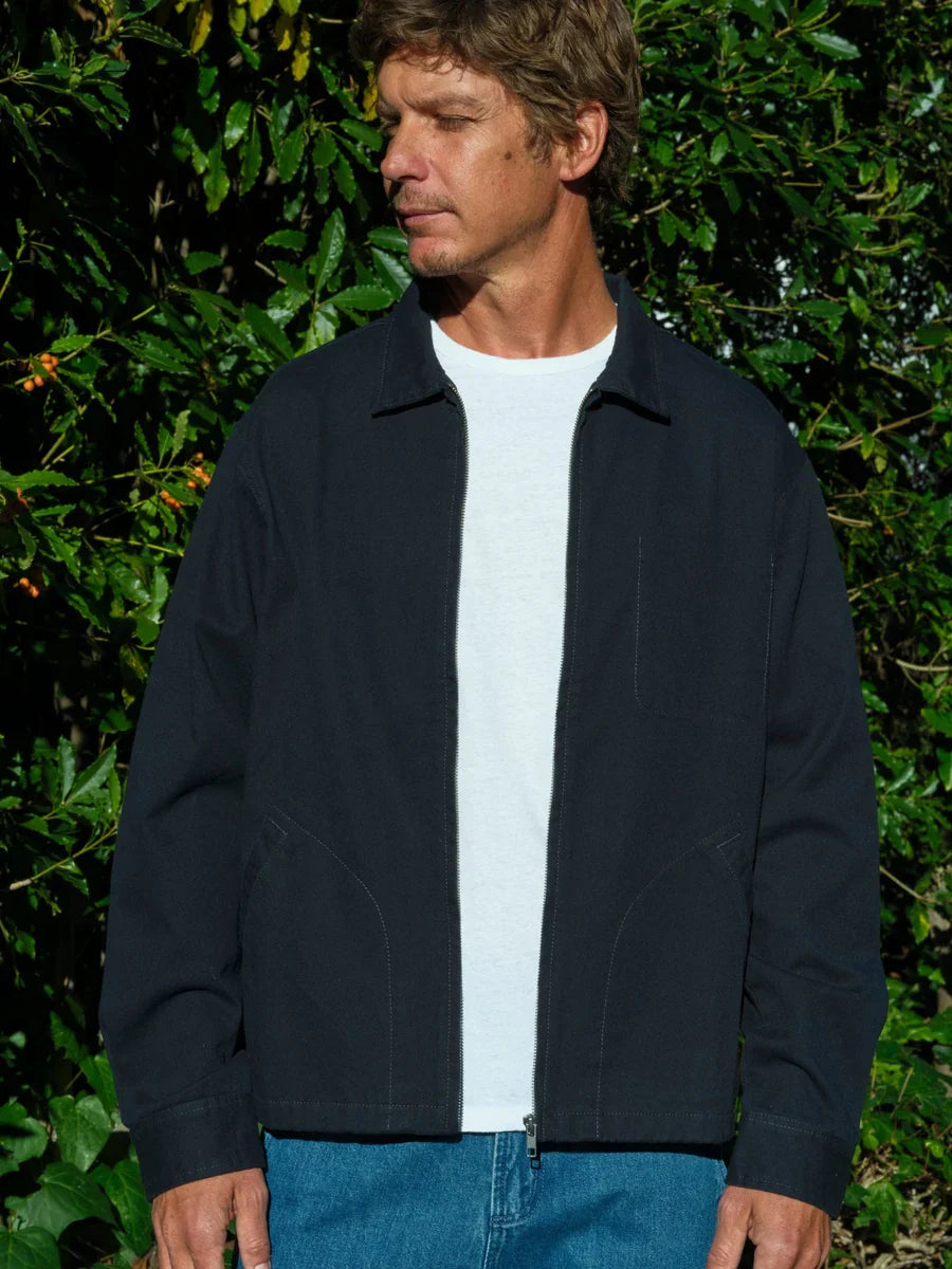 Three-quarter front view of a man wearing the Mollusk Gas Station Jacket in Navy open over a white tee, showing the spread collar, full-length zipper, and curved welt hand pockets in navy 100% cotton twill, styled with bright blue jeans against a sun-lit garden hedge
