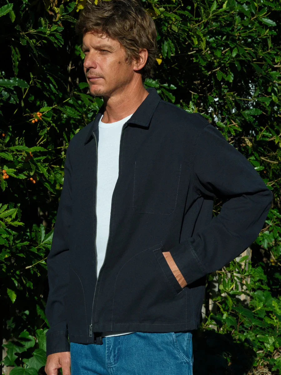 Side view of a man wearing the Mollusk Gas Station Jacket in Navy open with hand in welt pocket, showing the left chest interior pocket, relaxed silhouette, and navy 100% cotton twill fabric against a sun-lit garden hedge with red berries
