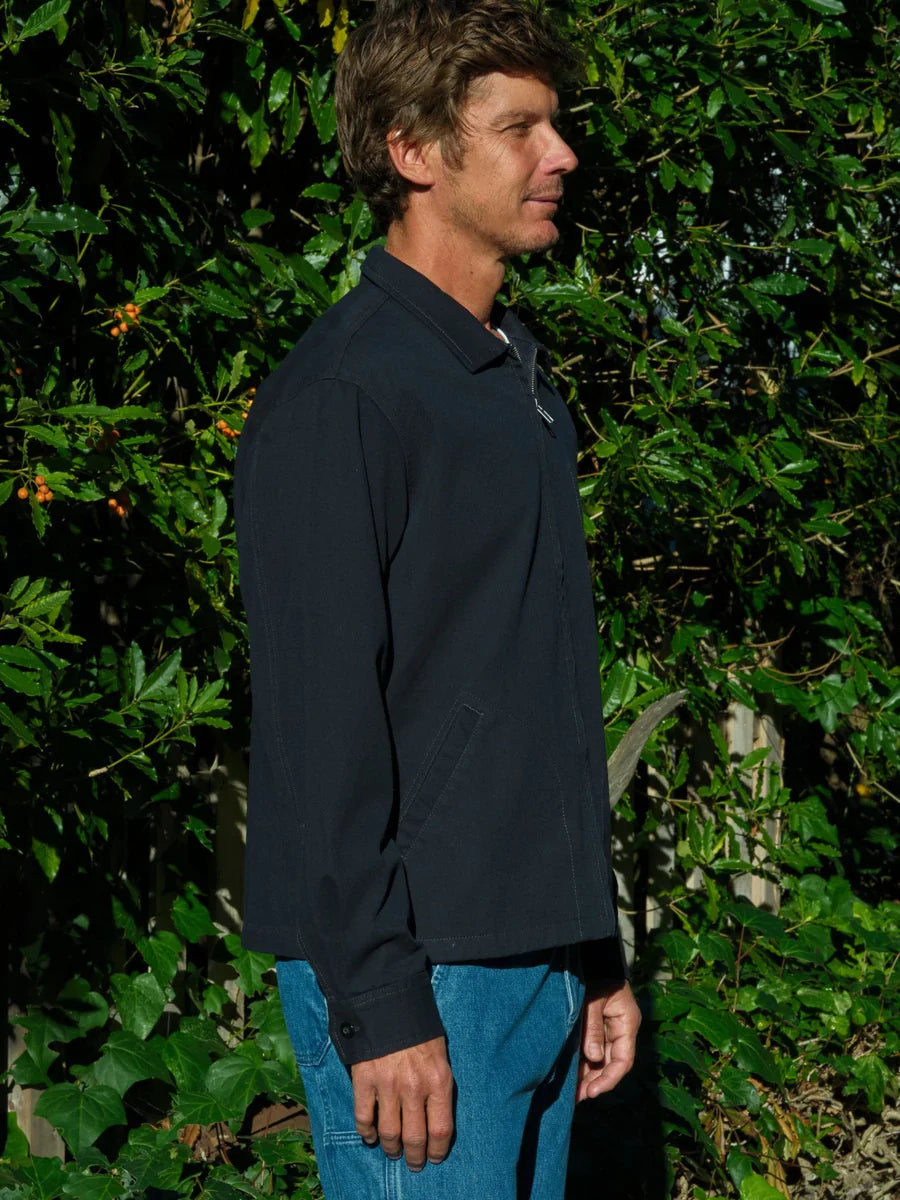 Pure side profile of a man wearing the Mollusk Gas Station Jacket in Navy zipped closed, showing the spread collar, full-length zipper pull, button cuff detail, and straight relaxed silhouette in navy 100% cotton twill, against a lush garden hedge
