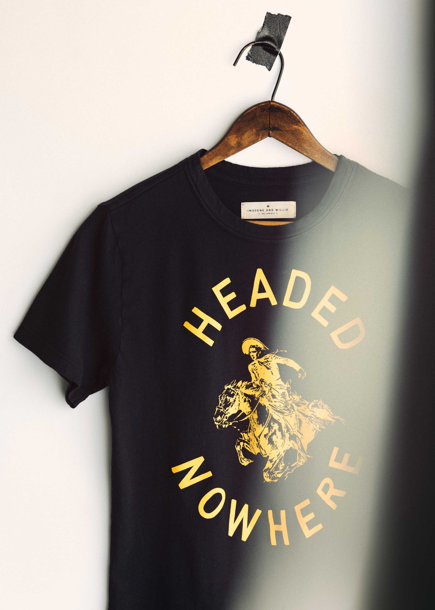 Angled close-up of The Headed Nowhere Tee by Imogene + Willie on a hanger, showing the Imogene and Willie label at the neckline and the gold screen-printed 'HEADED NOWHERE' circular graphic with cowboy on horseback on a black cotton tee