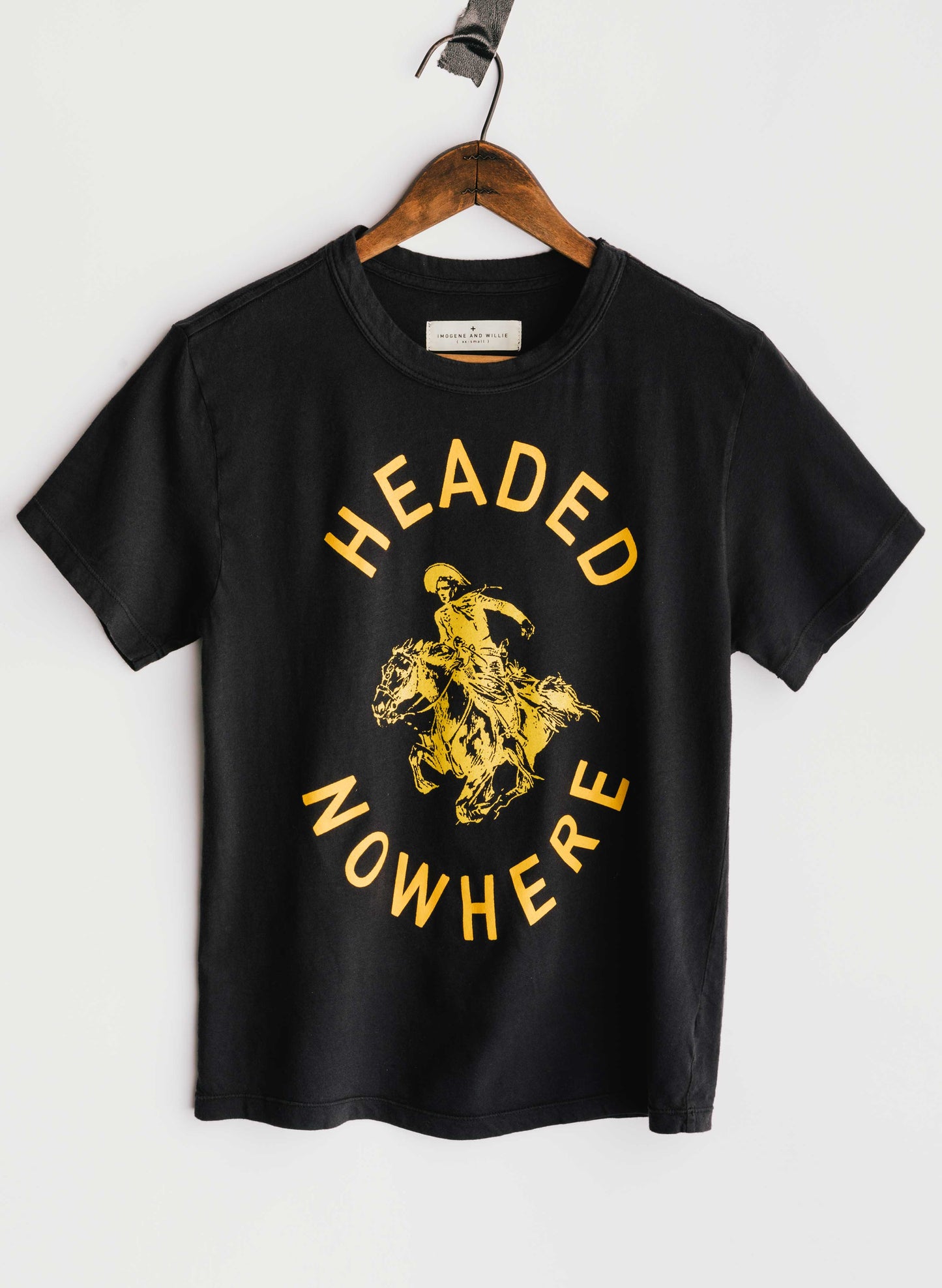 The Headed Nowhere Tee by Imogene + Willie on a wooden hanger, showing the full front with a circular screen-printed graphic reading 'HEADED NOWHERE' in gold vintage lettering around a gold illustration of a cowboy riding a rearing horse, on a black cotton tee