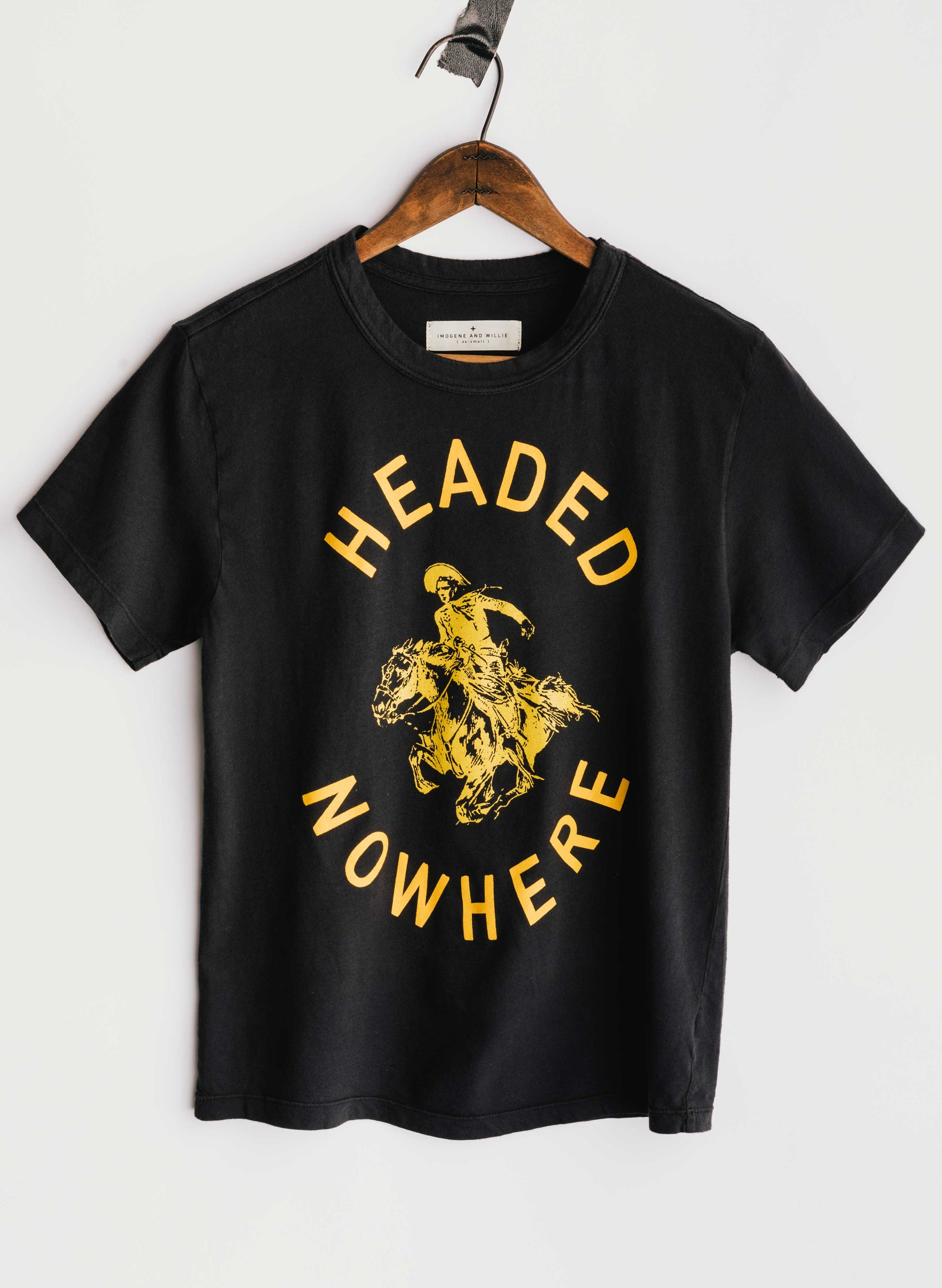 The Headed Nowhere Tee by Imogene + Willie on a wooden hanger, showing the full front with a circular screen-printed graphic reading 'HEADED NOWHERE' in gold vintage lettering around a gold illustration of a cowboy riding a rearing horse, on a black cotton tee
