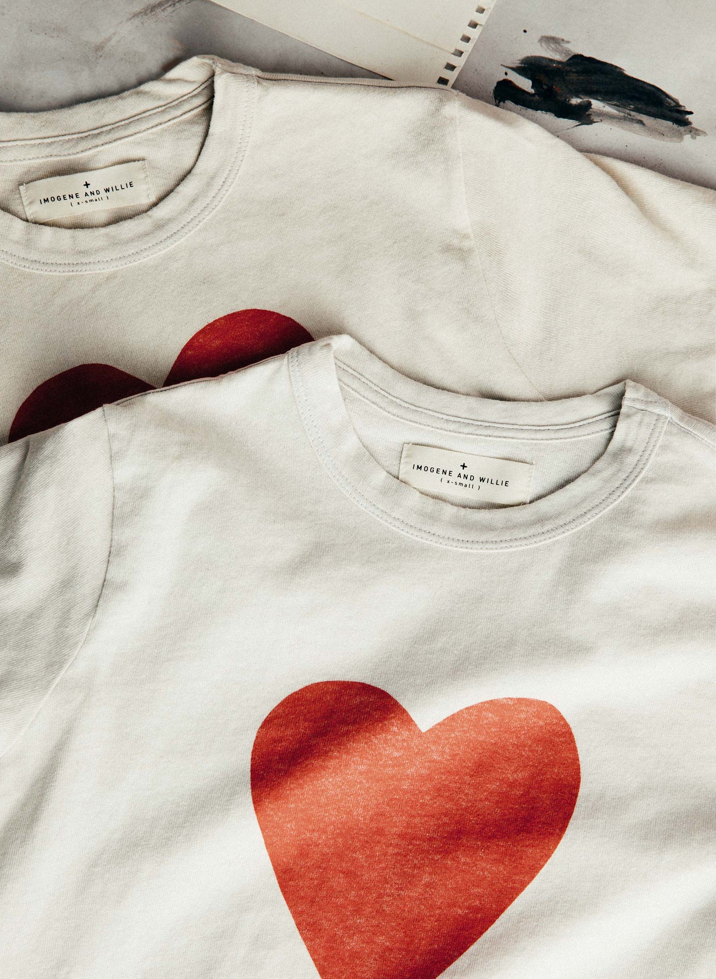Two tees stacked showing "Imogene + Willie" labels and heart print, with paint-brushed sketchbook in background