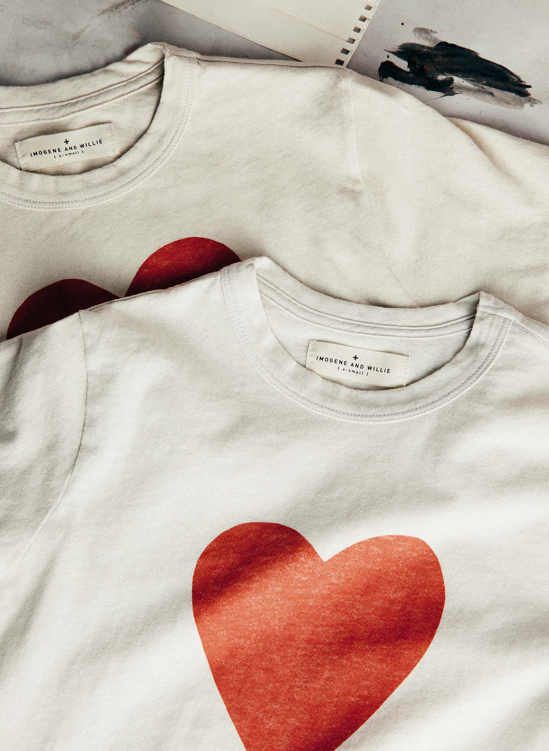 Two tees stacked showing "Imogene + Willie" labels and heart print, with paint-brushed sketchbook in background