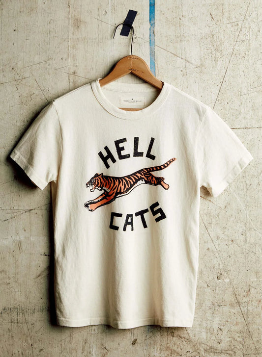 Cream short-sleeve tee on a wooden hanger with a vintage-style screen print of a leaping tiger and the text "Hell Cats," hung against a concrete wall.
