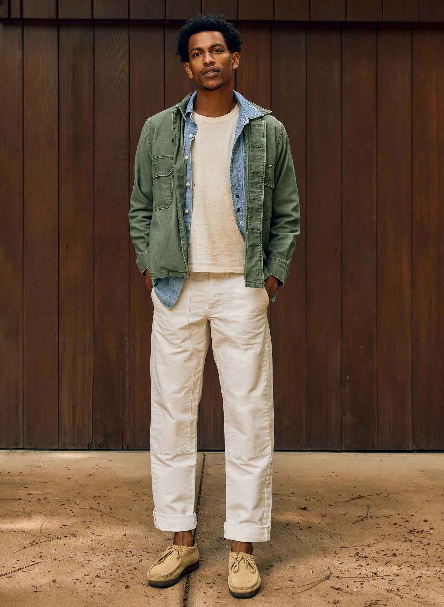 Man wearing the Hencye Canvas Workpant in Natural with a cuffed hem, cream tee, and green field jacket, standing in front of a wood-paneled wall with hands in pockets
