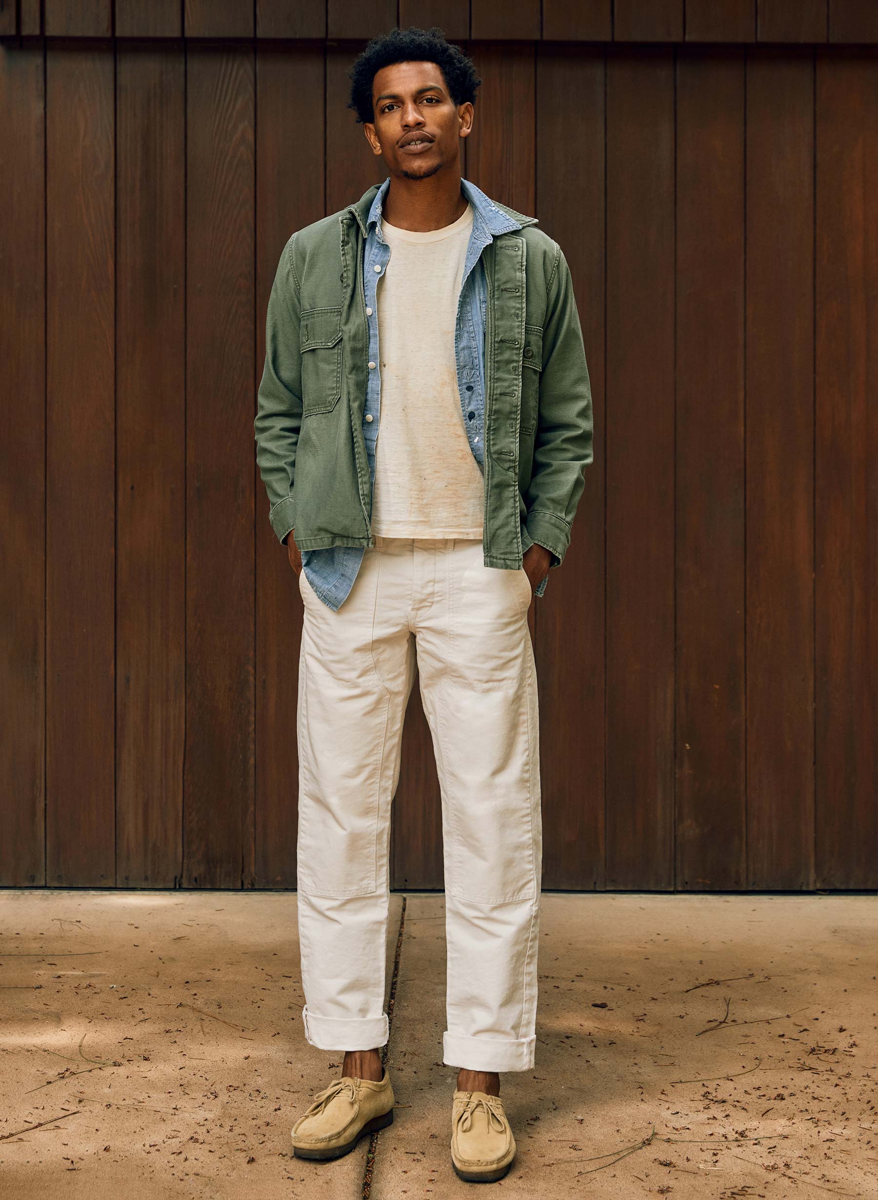 Man wearing the Hencye Canvas Workpant in Natural with a cuffed hem, cream tee, and green field jacket, standing in front of a wood-paneled wall with hands in pockets