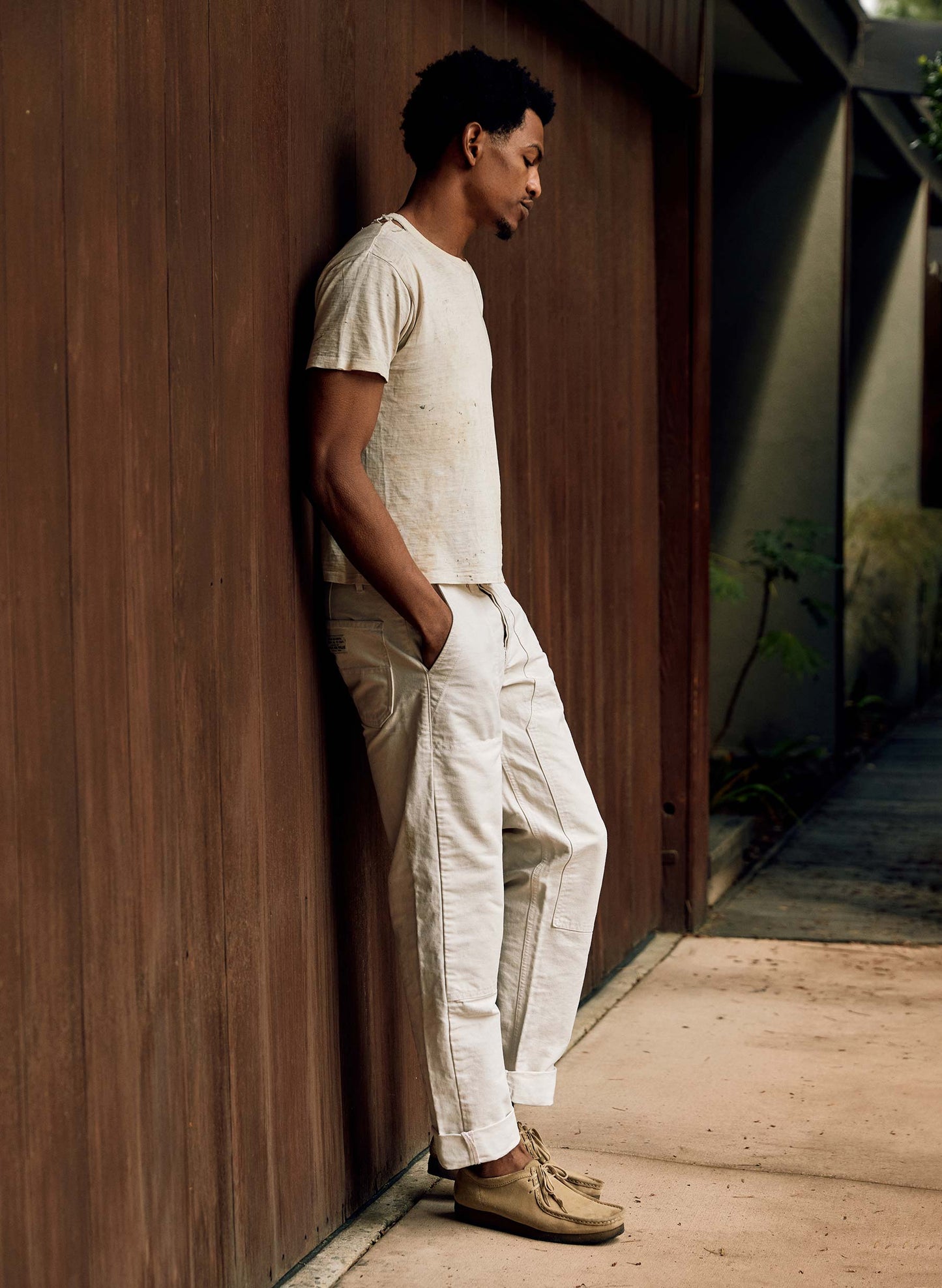 Side profile of a man leaning against a wood-paneled wall wearing the Hencye Canvas Workpant in Natural with a cuffed hem, cream tee, and moccasin shoes, showing the straight leg silhouette