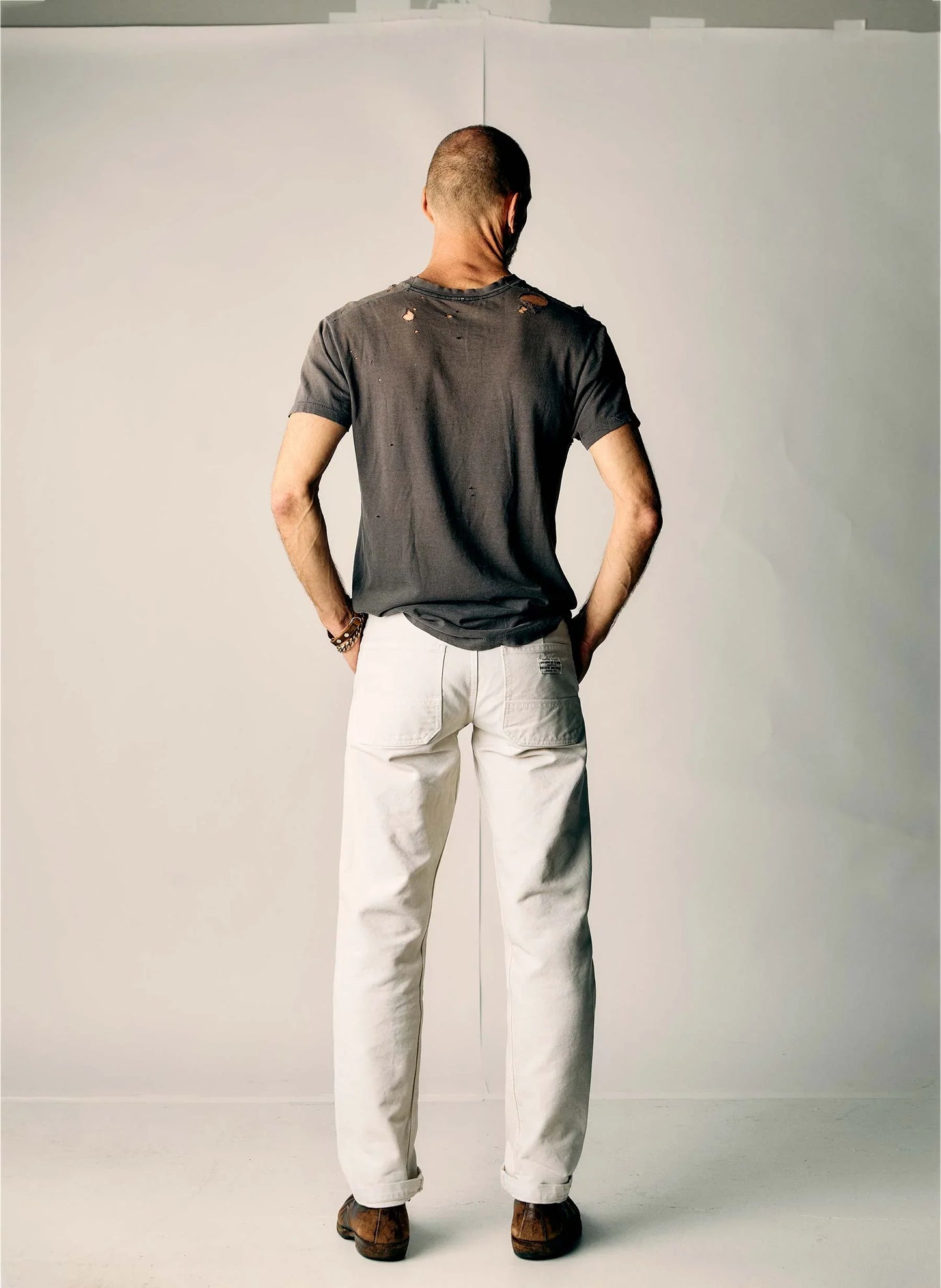 Full back view of a man wearing the Hencye Canvas Workpant in Natural with a cuffed hem and charcoal tee, showing the relaxed straight leg fit and overall silhouette