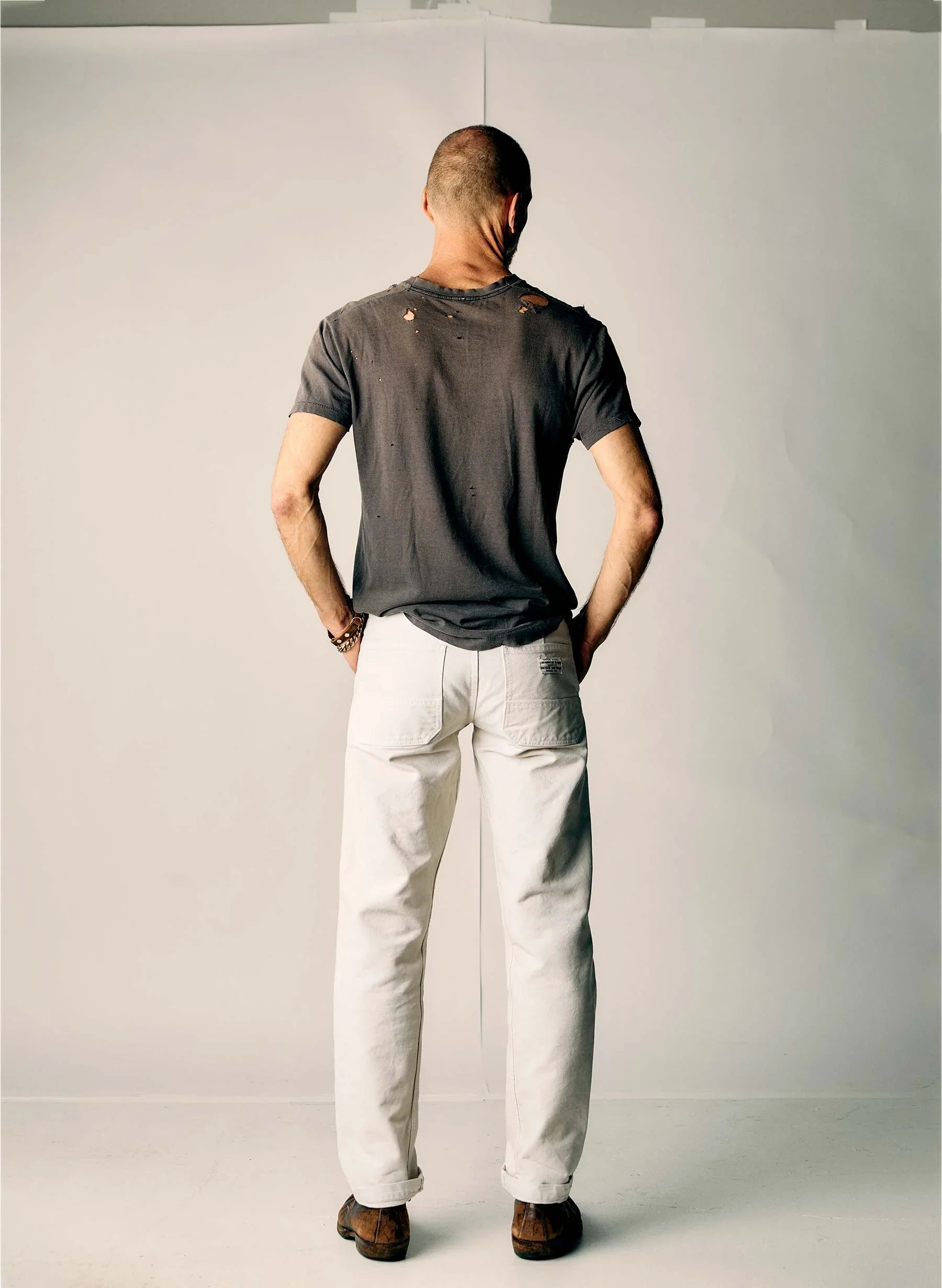 Full back view of a man wearing the Hencye Canvas Workpant in Natural with a cuffed hem and charcoal tee, showing the relaxed straight leg fit and overall silhouette