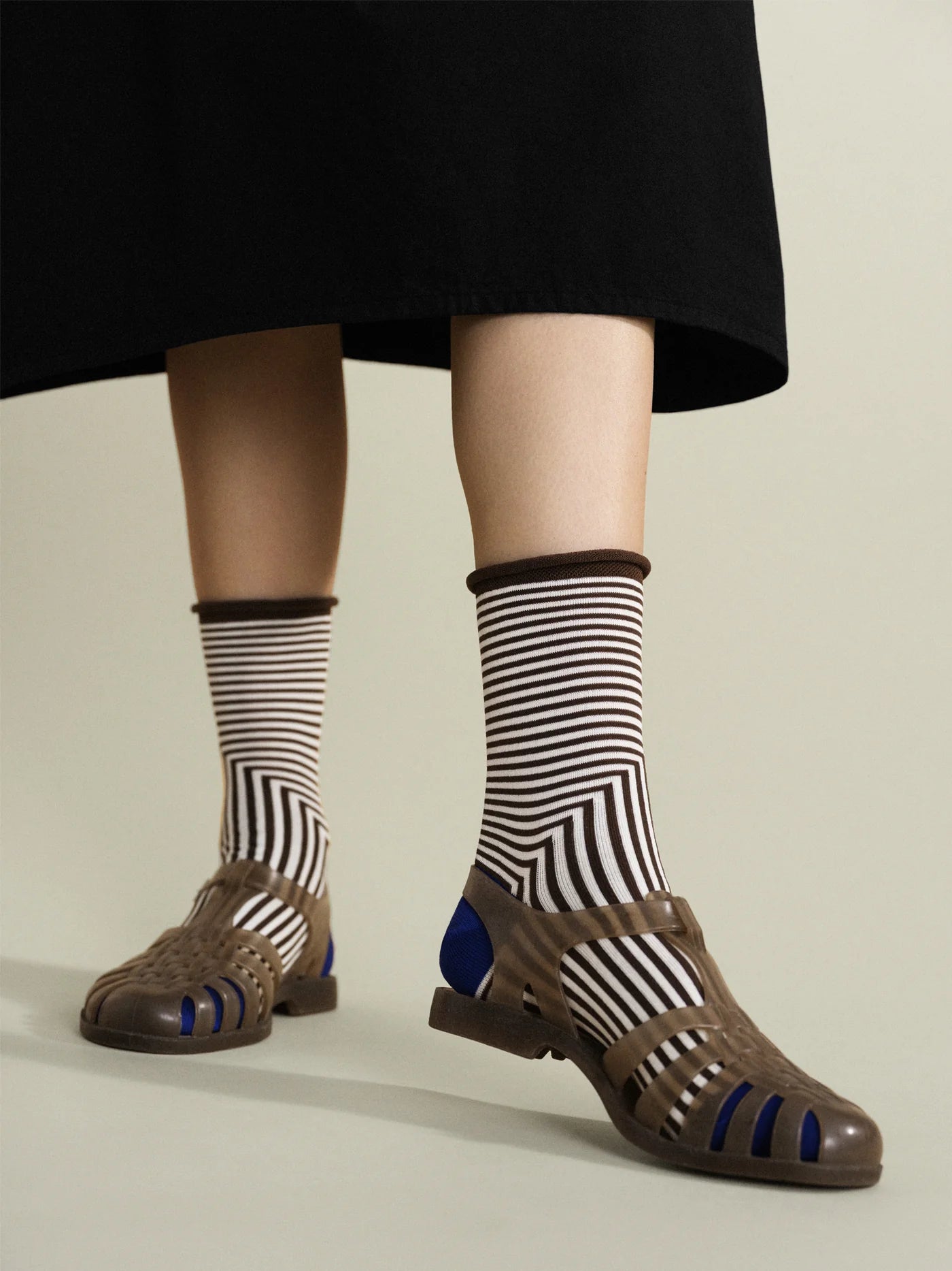 Corbusier Crew socks in Brown — graphic op-art stripe with brown rolled cuff and cobalt blue heel, worn with olive jelly sandals under a black midi skirt

