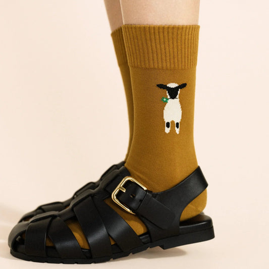 Nice to See Ewe Crew socks in Ochre — warm golden ochre crew with lamb motif and ribbed cuff, worn with black leather buckle sandals on a cream background
