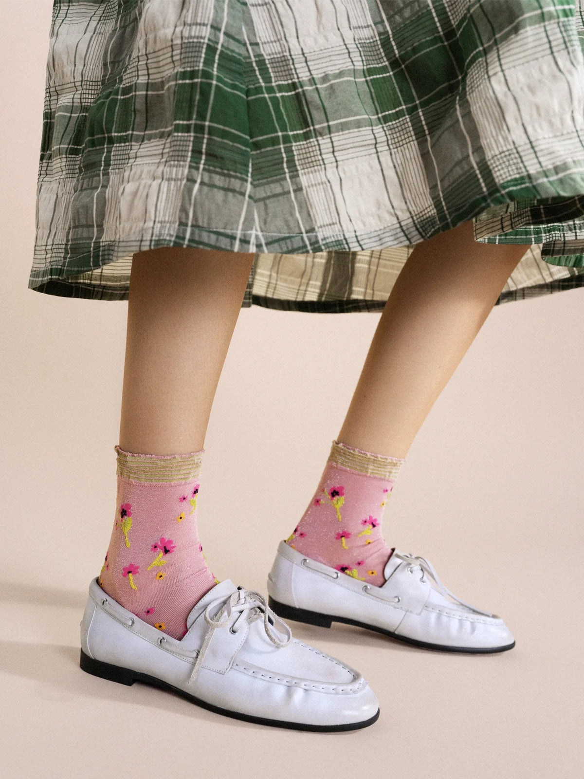 Lumiesta Floral Short Crew socks in Pink — sheer pink nylon short crew with allover floral motif and striped ruffle cuff, worn with white leather boat shoes under a green plaid midi skirt on a cream background
