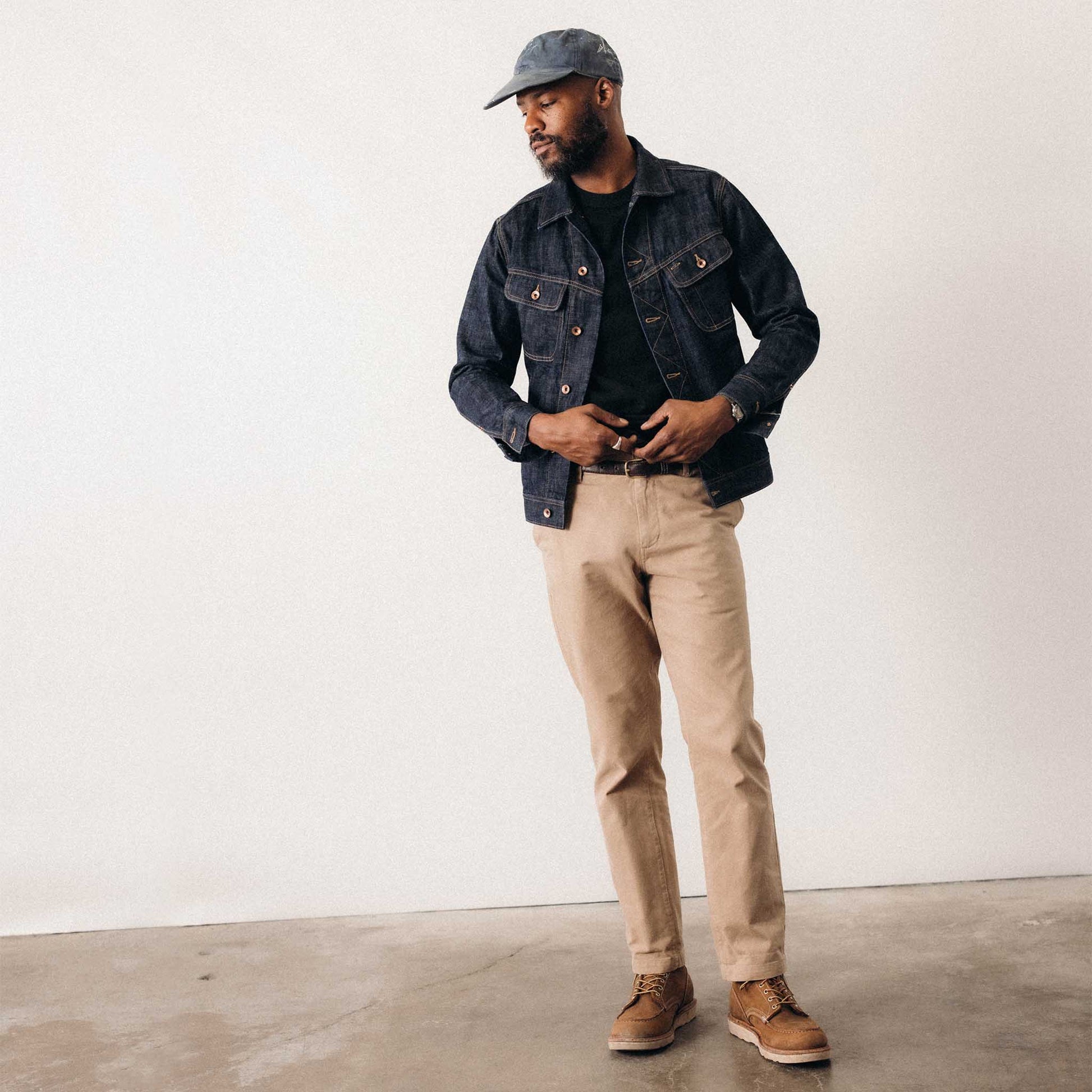 Full-length shot of a man wearing The Long Haul Jacket in Rinsed Organic Selvedge by Taylor Stitch, styled with khaki pants, brown boots, and a grey cap against a white studio wall