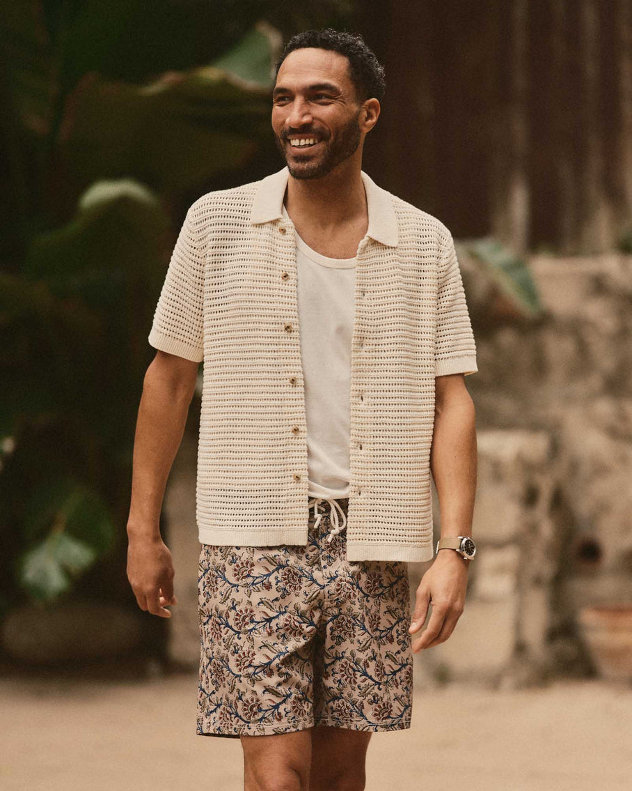 Man wearing a beige textured shirt over a white t-shirt and floral shorts, standing outdoors.