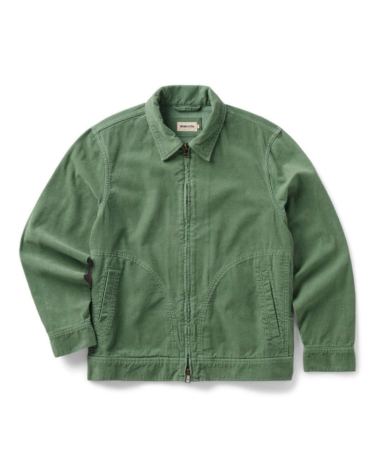 Flat lay of the Taylor Stitch Piston Jacket in Ivy Corduroy on a white background, showing the full jacket from the front. The jacket is a muted sage green in wide-wale organic cotton corduroy with a point collar, a YKK two-way separating zipper down the center front, two curved lower hand warmer pockets, long sleeves with single-button cuffs, and a ribbed hem. A small woven Taylor Stitch label is visible at the collar.
