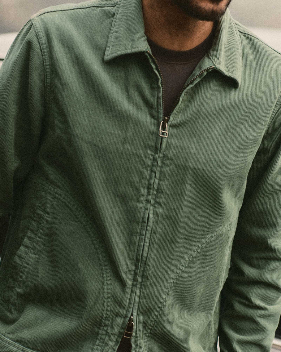 Close-up on-body shot of a man wearing the Taylor Stitch Piston Jacket in Ivy Corduroy, cropped from chin to waist against a soft gray background. The wide-wale corduroy texture is clearly visible across the chest and shoulders. The YKK zipper is half-open, revealing a dark navy crewneck underneath. The jacket's point collar lies flat and the curved pocket opening is partially visible at lower left.
