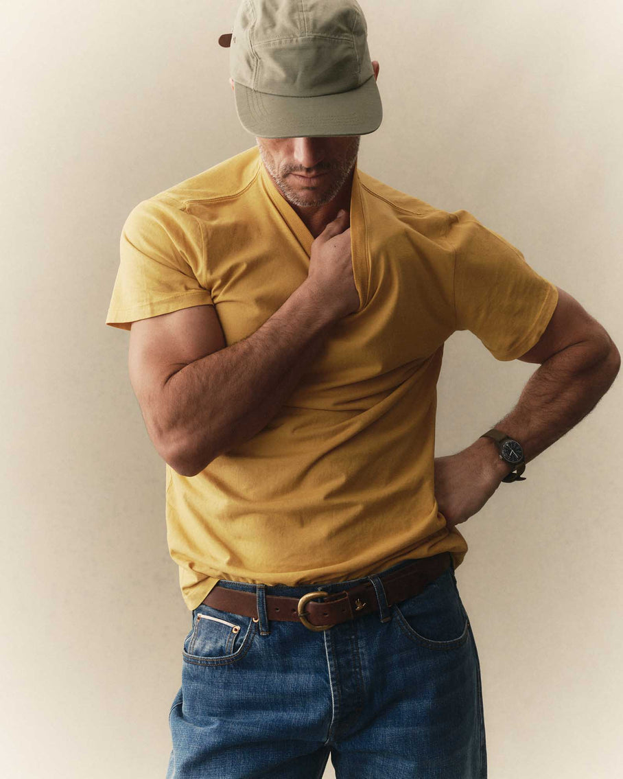 Man wearing Taylor Stitch Cotton Hemp Tee in Goldfinch yellow with blue jeans and olive cap, hand at collar