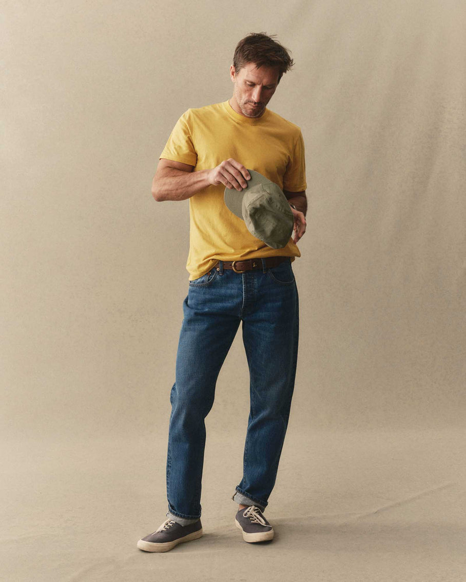 Man wearing Taylor Stitch Cotton Hemp Tee in Goldfinch yellow with blue jeans and sneakers, holding an olive hat