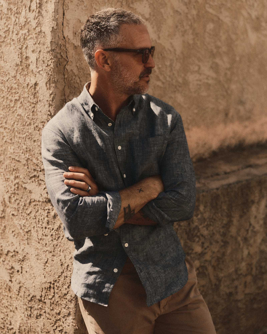 Model wearing Taylor Stitch The Jack in Ocean Linen, side profile arms crossed against a textured adobe wall, showing ocean blue-grey washed European linen, button-down collar, and tailored pleat-free silhouette, styled with tan chinos and sunglasses
