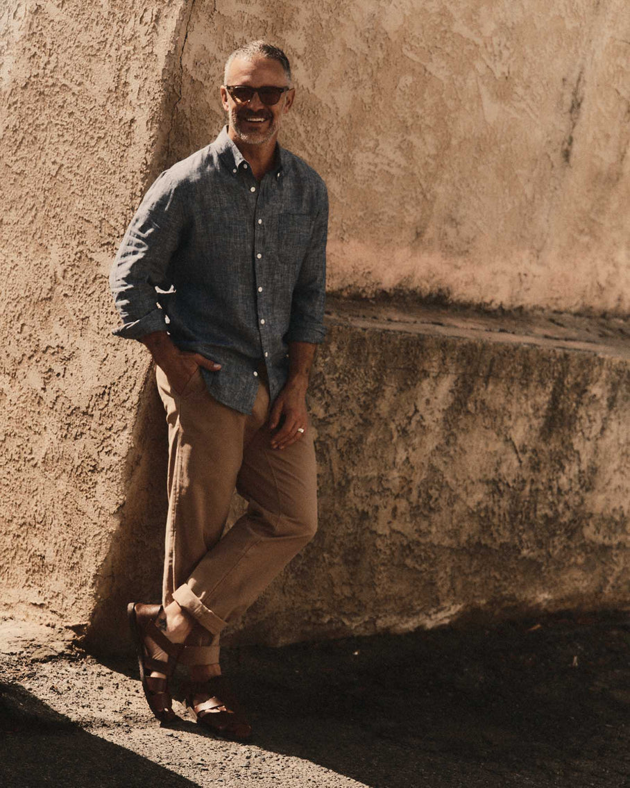 Model wearing Taylor Stitch The Jack in Ocean Linen, full-length front view leaning against an adobe wall outdoors, showing ocean blue-grey washed European linen, button-down collar, single chest pocket, and tailored silhouette, styled with tan chinos and leather sandals
