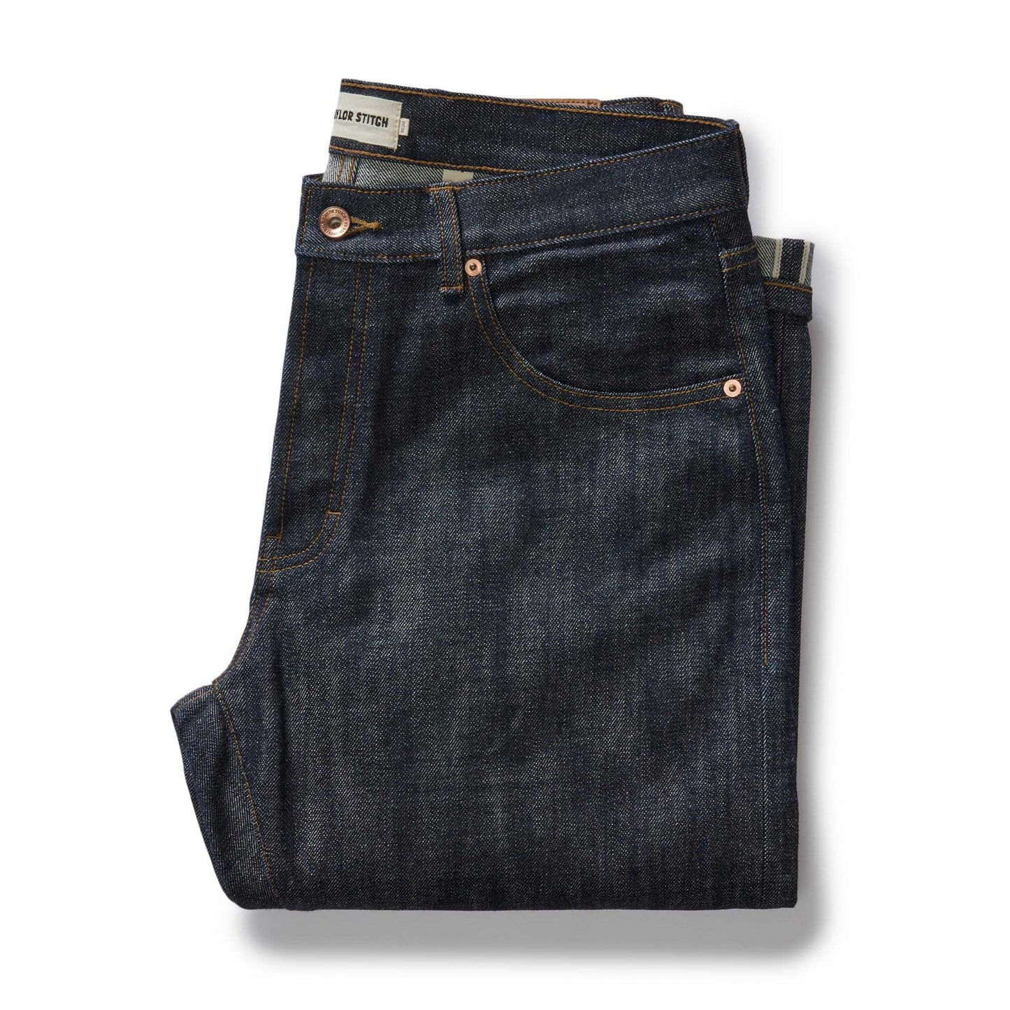 Folded pair of The Democratic Jean in Rigid Organic Selvedge by Taylor Stitch, showing the dark indigo raw denim, copper rivets, and Taylor Stitch waistband label on a white background