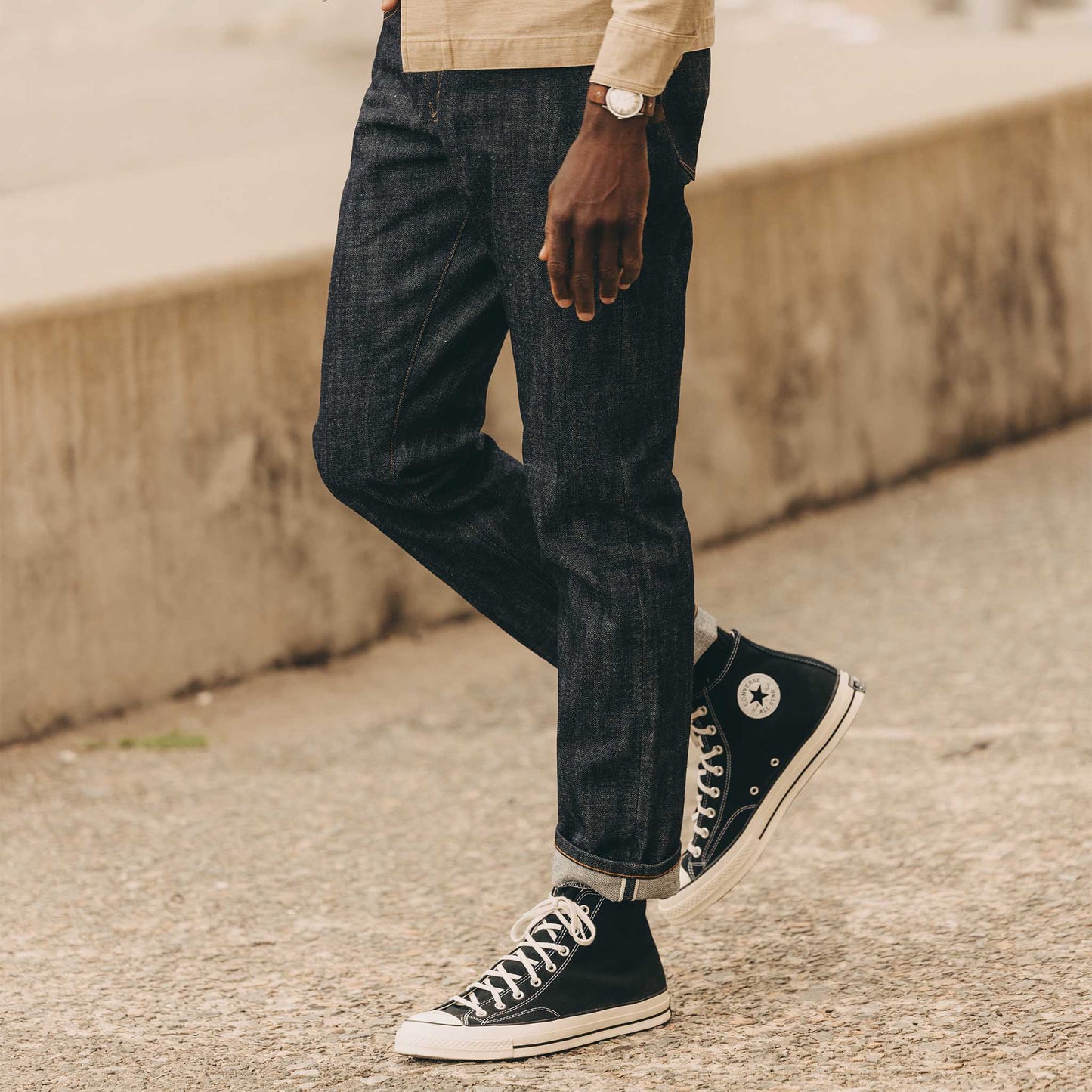 Man wearing The Democratic Jean in Rigid Organic Selvedge by Taylor Stitch, styled with a tan overshirt and black Converse high-tops, showing the straight leg fit and cuffed selvedge hem