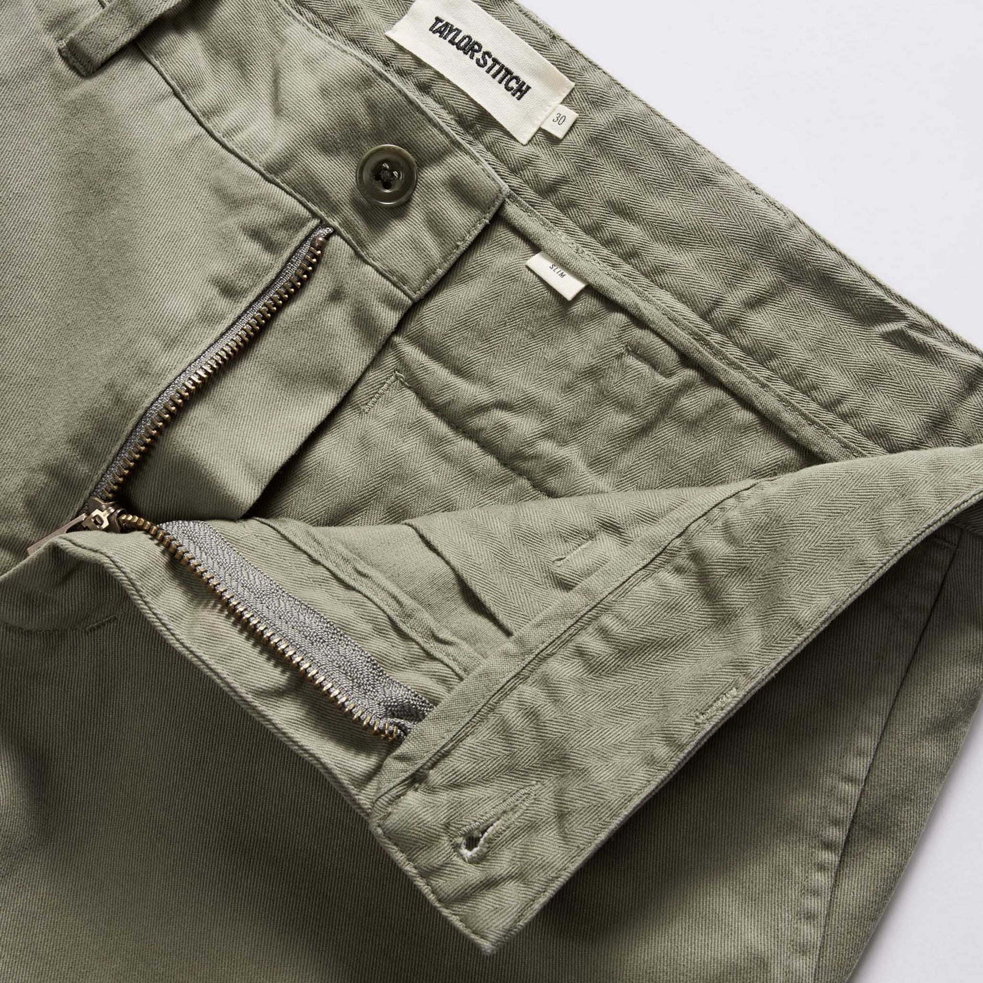 Close-up of olive green pants showing the waistband, button closure, zippered pocket, and interior pocket lining. A sewn-in label reads TAYLOR STITCH.