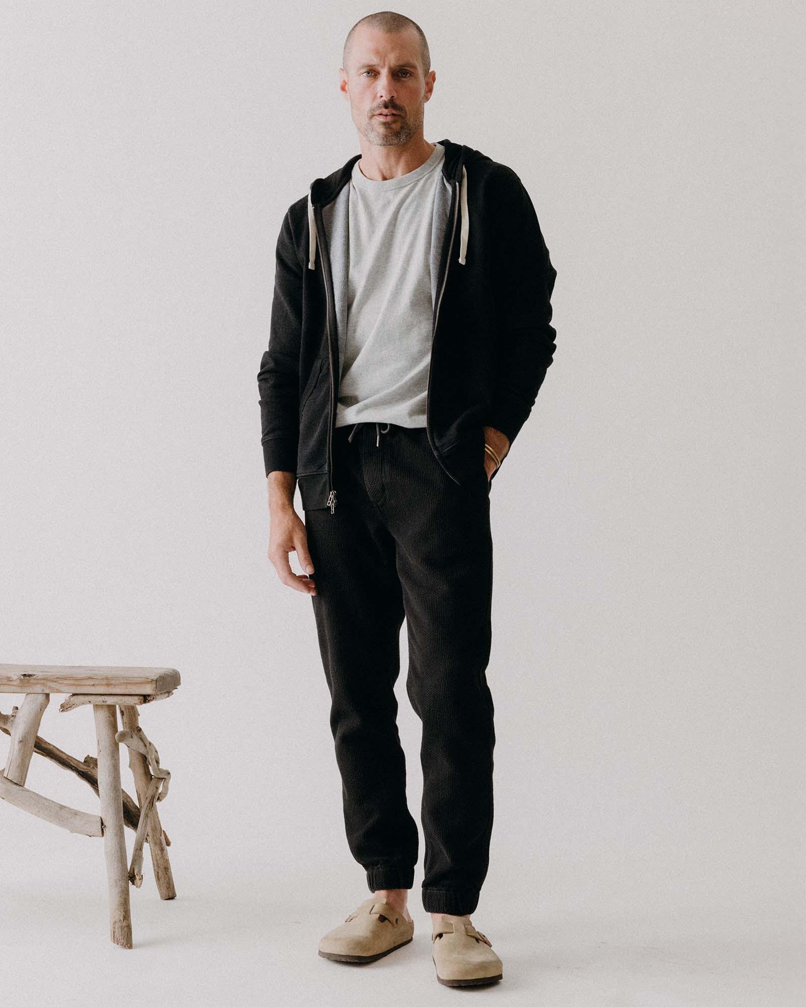 Man wearing a black jacket, gray shirt, and black pants standing against a white background.