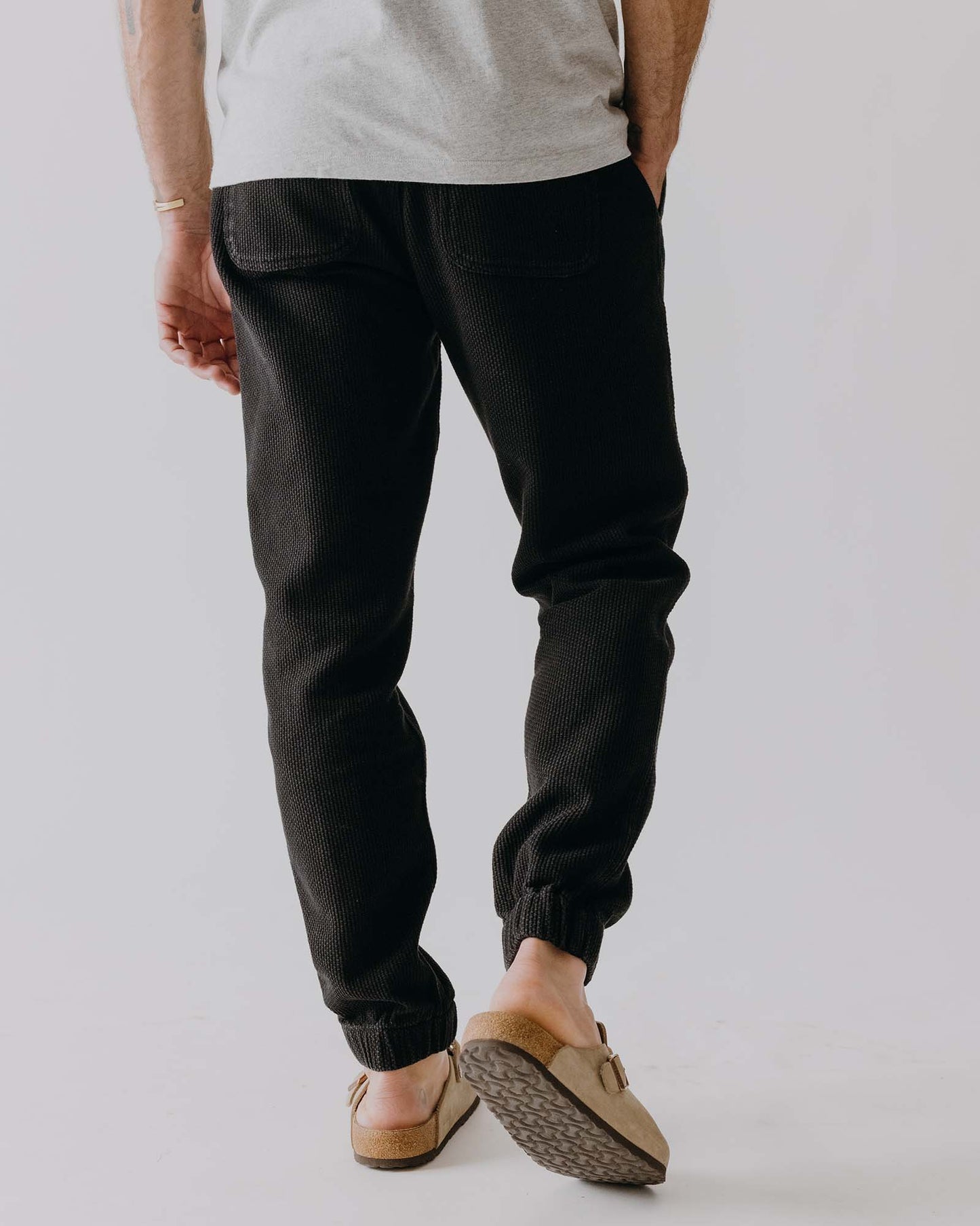 Person wearing black pants and beige shoes on a plain background, shown from the back
