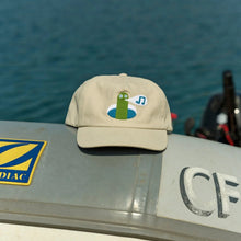 Load image into Gallery viewer, Beige cap with a green character on a boat with water in the background
