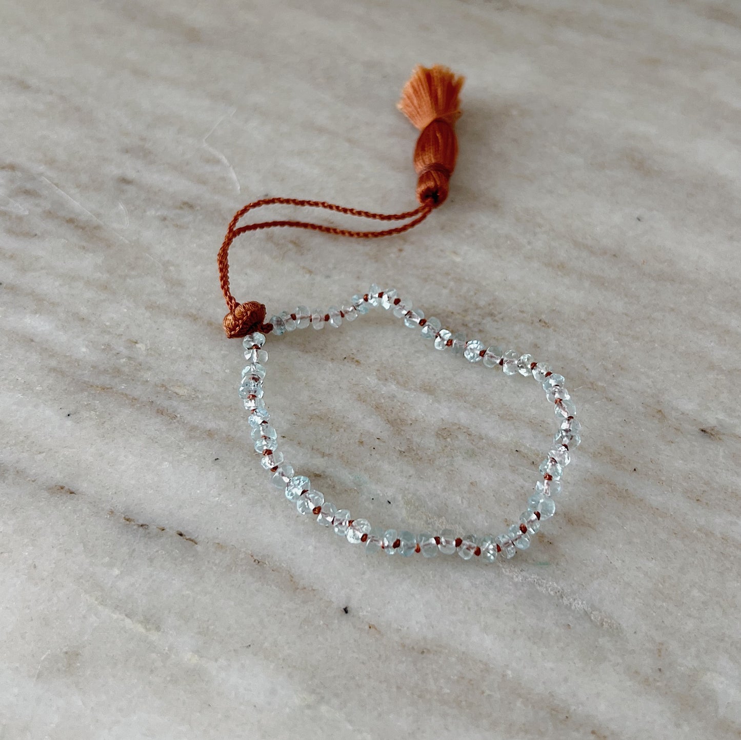 An aquamarine beaded bracelet with a waxed silk cord and an adjustable tassel closure, displayed on a grey marble slab.