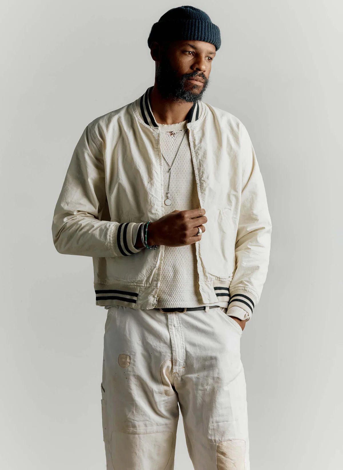 Man wearing The Madisonville Jacket by Imogene + Willie open over a cream mesh tee and white work pants, with a navy beanie and layered necklaces, showing the relaxed fit and black and white striped rib trim