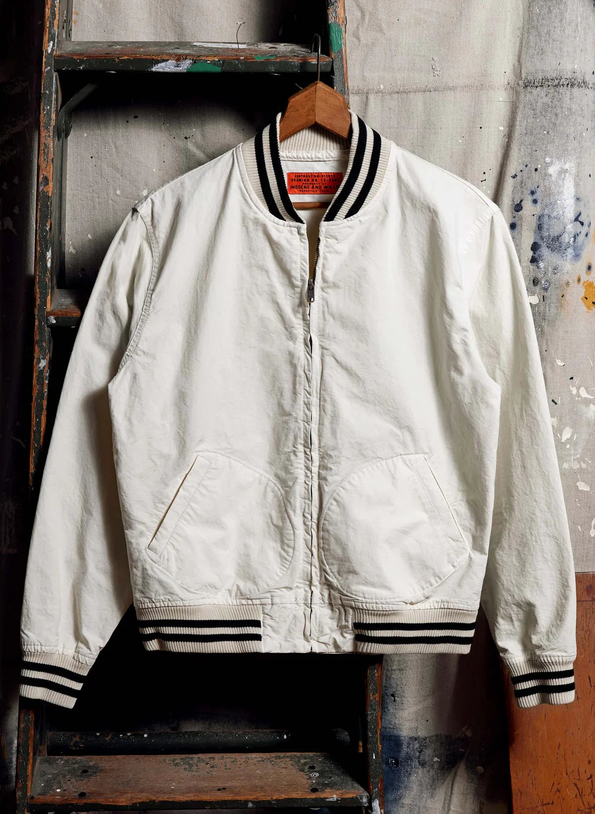 The Madisonville Jacket by Imogene + Willie on a wooden hanger in a workshop setting, showing the full front in natural off-white 8 oz cotton with black and white striped rib collar, cuffs, and hem, antique brass zipper, and slanted welt pockets