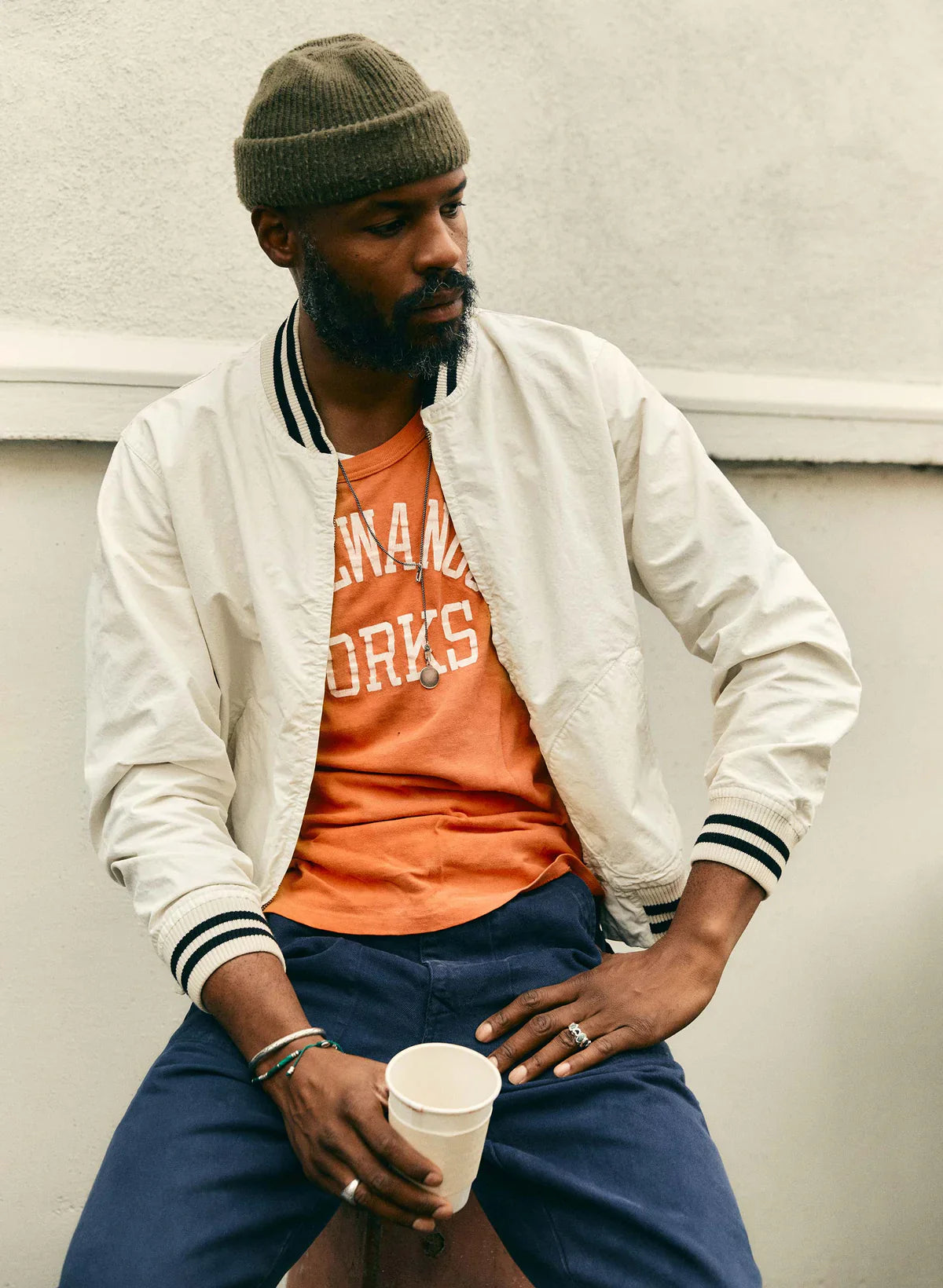 Man seated wearing The Madisonville Jacket by Imogene + Willie open over an orange graphic tee and navy pants, holding a cup, with an olive beanie, showing the jacket's relaxed fit and striped rib cuffs