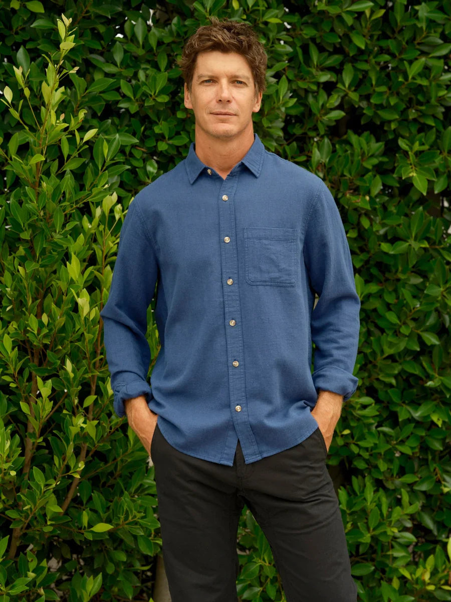 Model wearing Mollusk Surf Shop One Pocket Shirt in Indigo Flannel, full front view against lush green hedge showing medium indigo blue 80% cotton 20% linen flannel, point collar, long sleeves, tonal corozo-style button-front placket, and single left-chest patch pocket, styled with black trousers, hands in pockets
