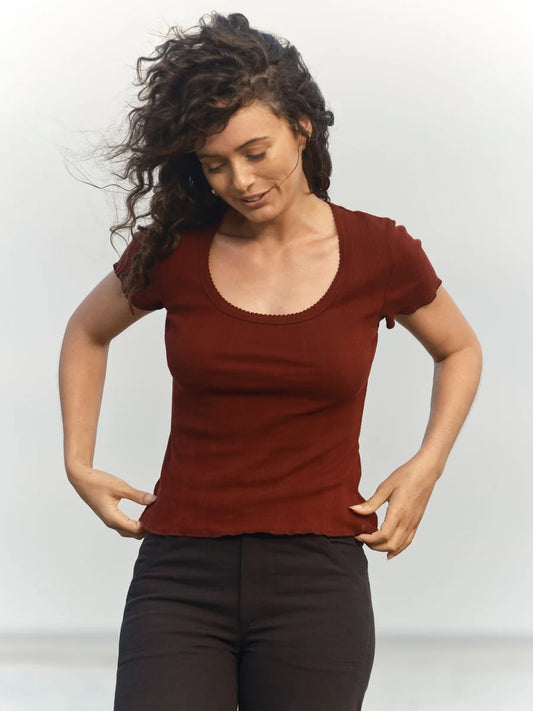 Female model wearing the Mollusk Surf Shop Pointelle Tee in Malbec, a deep red-brown short-sleeve cotton tee with scoop neck and ruffled trim, styled with black pants against a light grey background
