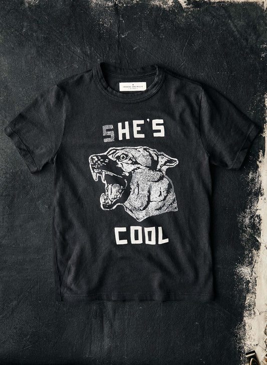 The She's Cool Tee by Imogene + Willie laid flat on a dark textured surface, showing the full front with a vintage-style screen-printed graphic of a snarling panther head and the text 'SHE'S COOL' in white distressed lettering on a black cotton tee