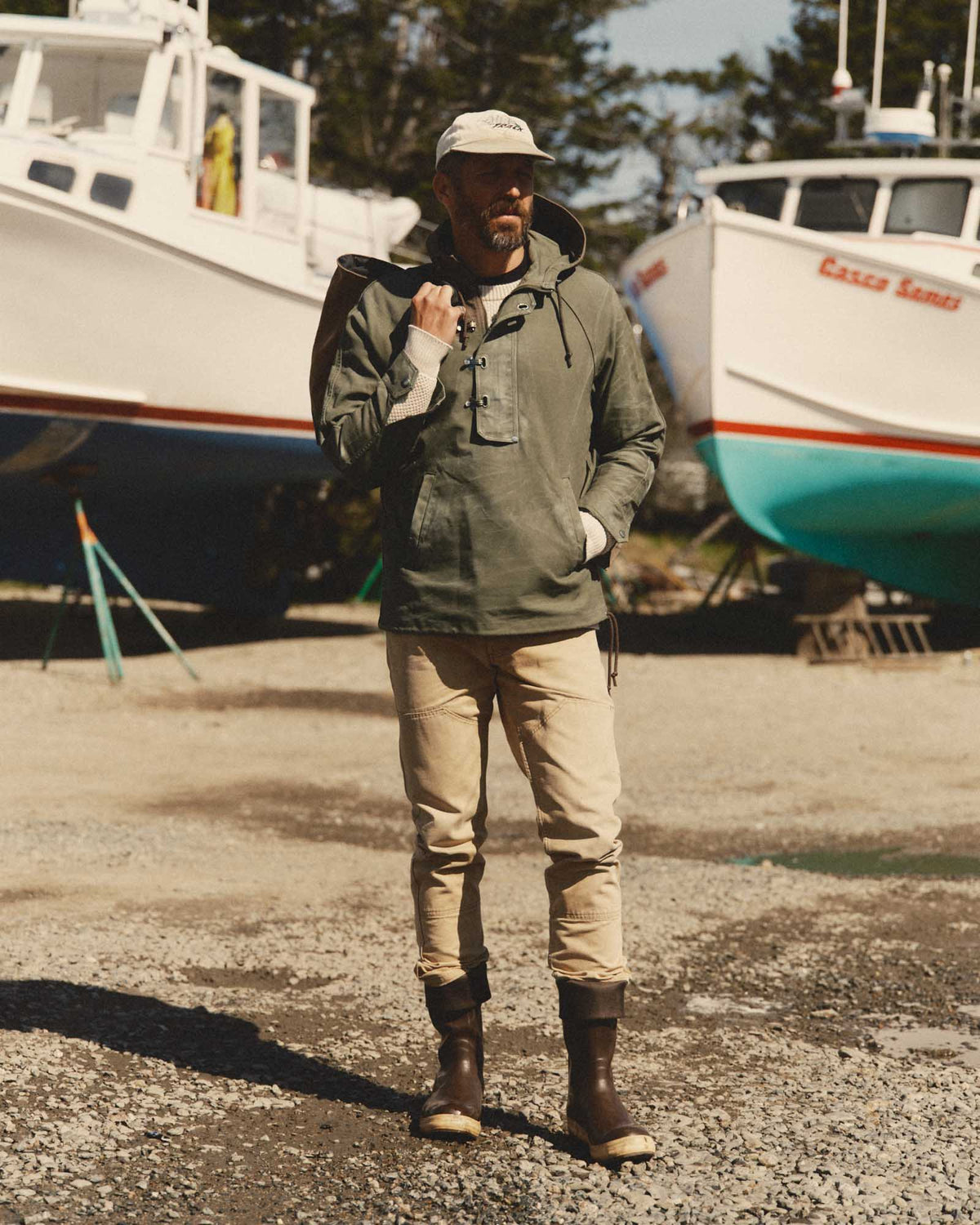Full-length view of a model wearing the fatigue olive Hatteras Anorak with tan pants and boots in a boatyard.
