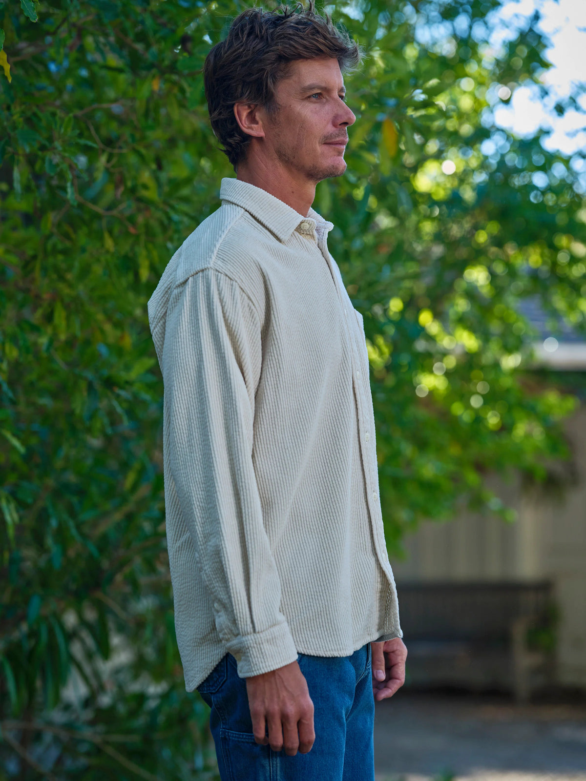 Side profile of a man standing outdoors wearing the Mollusk Thurston Shirt in Natural Corduroy, showing the relaxed boxy silhouette, slightly dropped shoulders, long sleeves, and wide bubble corduroy wale texture in cream-natural 100% cotton, styled with medium blue jeans against a soft-focus green garden backdrop
