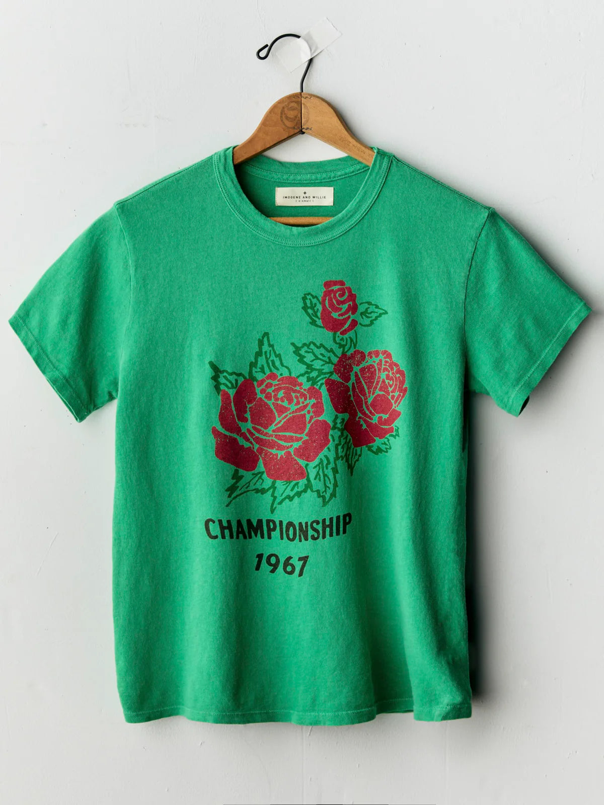 A green short-sleeve t-shirt hanging on a wooden hanger against a light gray wall. The shirt features a graphic print of three red roses with green leaves and the text 'CHAMPIONSHIP 1967' in black below the roses.