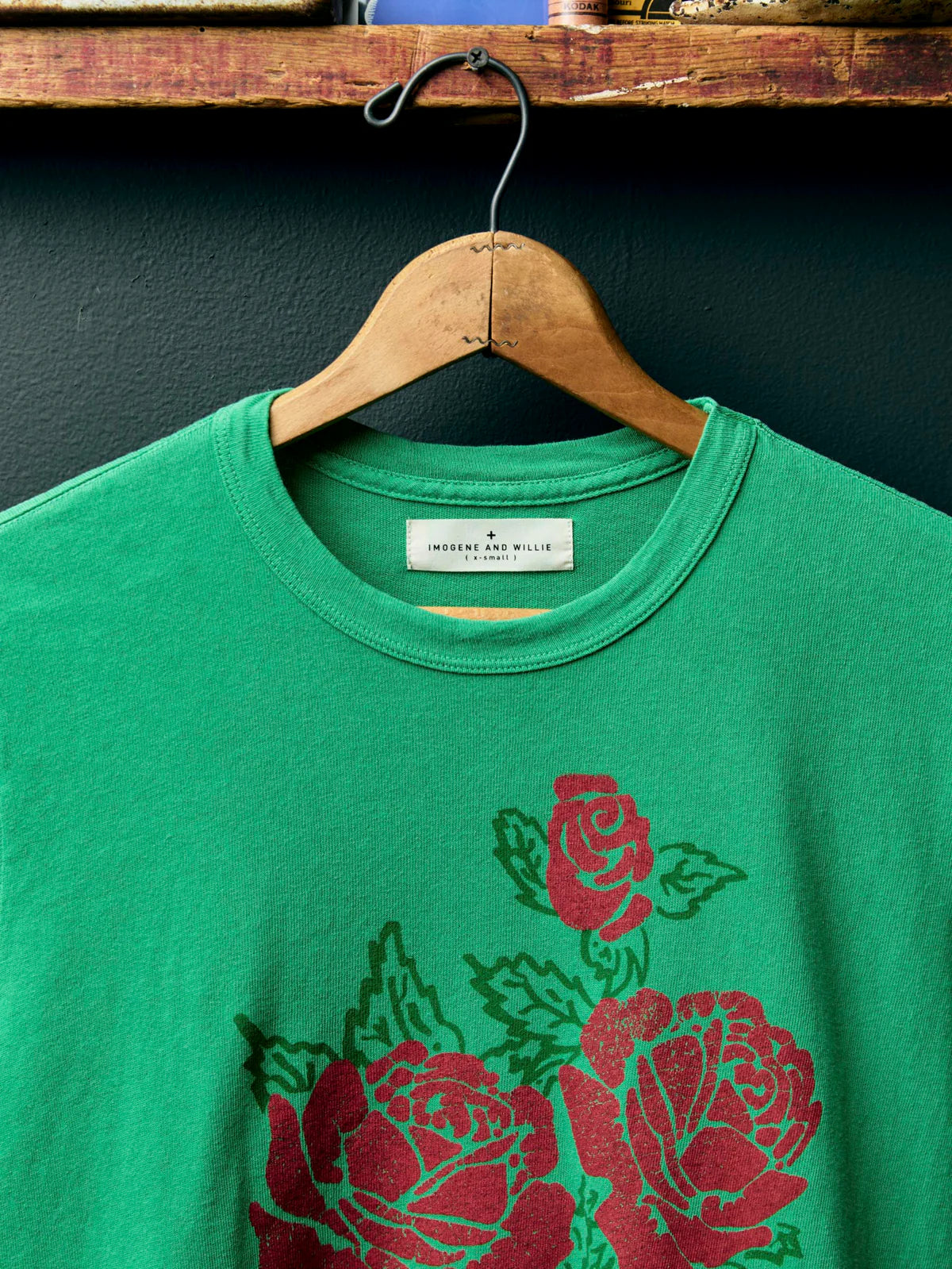 A close up shot of green crewneck t-shirt hanging on a wooden hanger against a dark textured wall. The shirt features a graphic print of red roses with green leaves on the front.