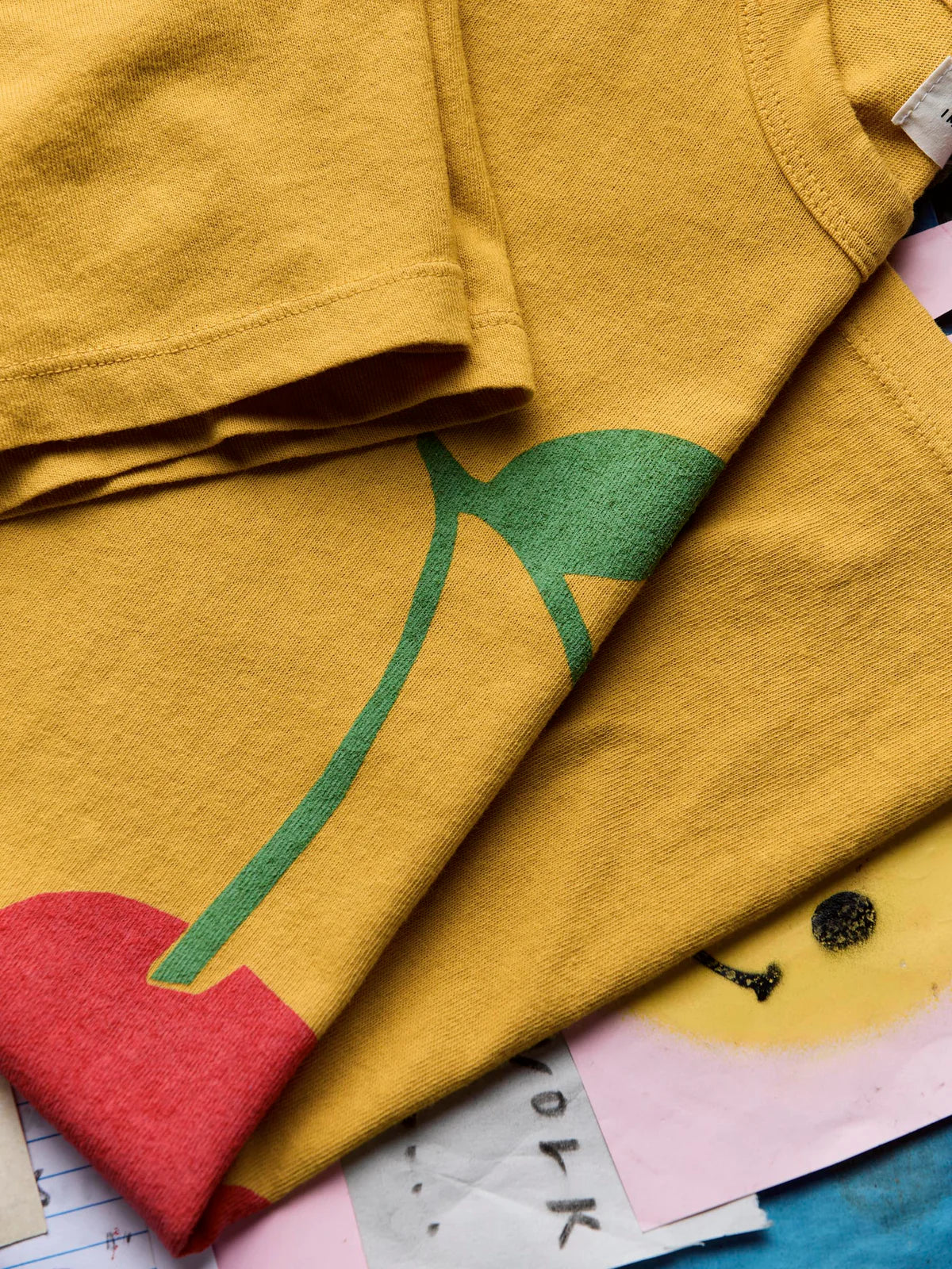 Flat lay close-up of The Cherry Tee in Gold showing the screen-printed cherry stem and leaf graphic, highlighting the hand-drawn artwork detail and gold cotton fabric texture