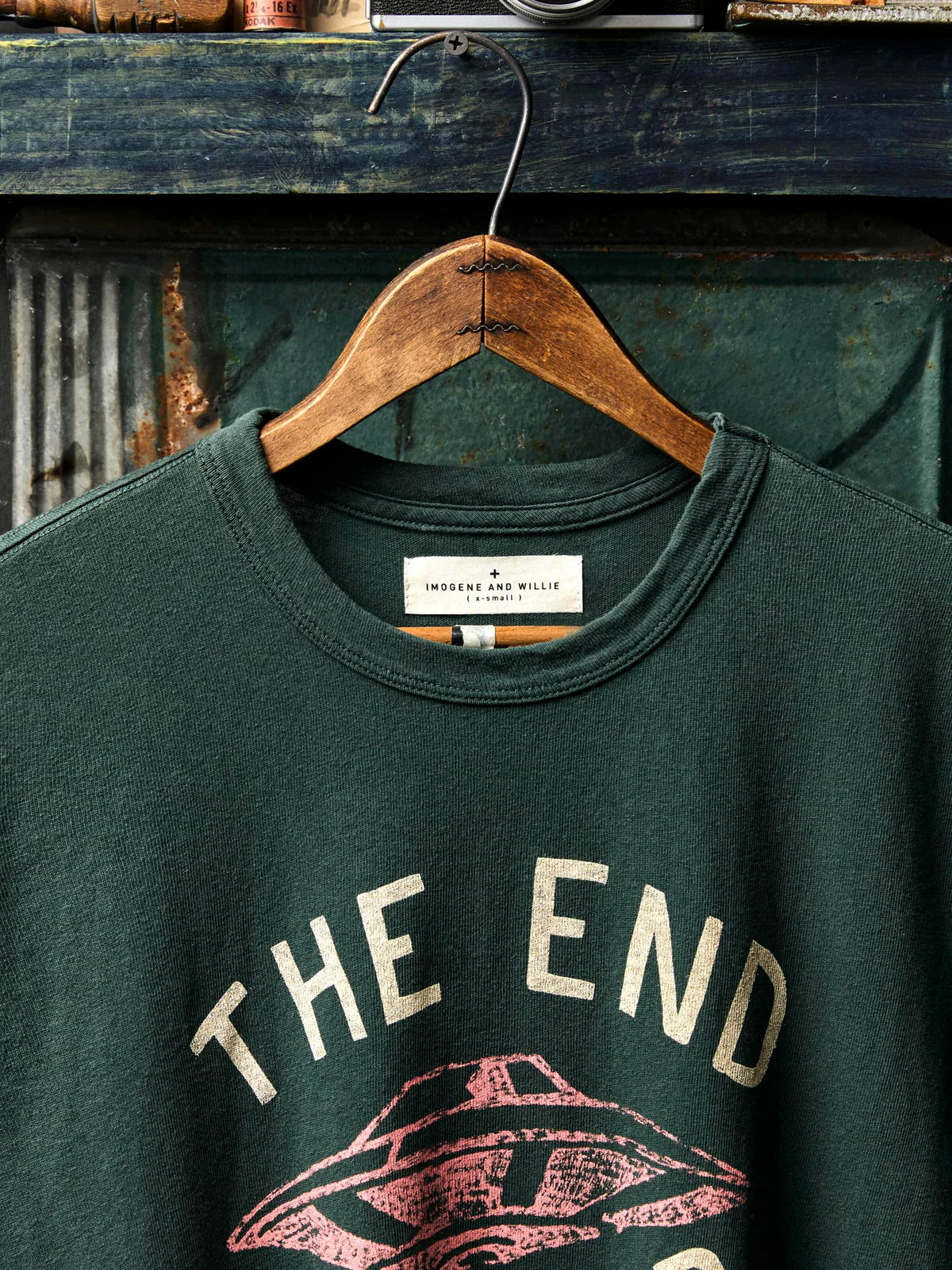 Close-up of the Imogene + Willie label on the neck of a dark spruce green tee, with a faded "The End Is Near" screen print and UFO graphic visible below.
